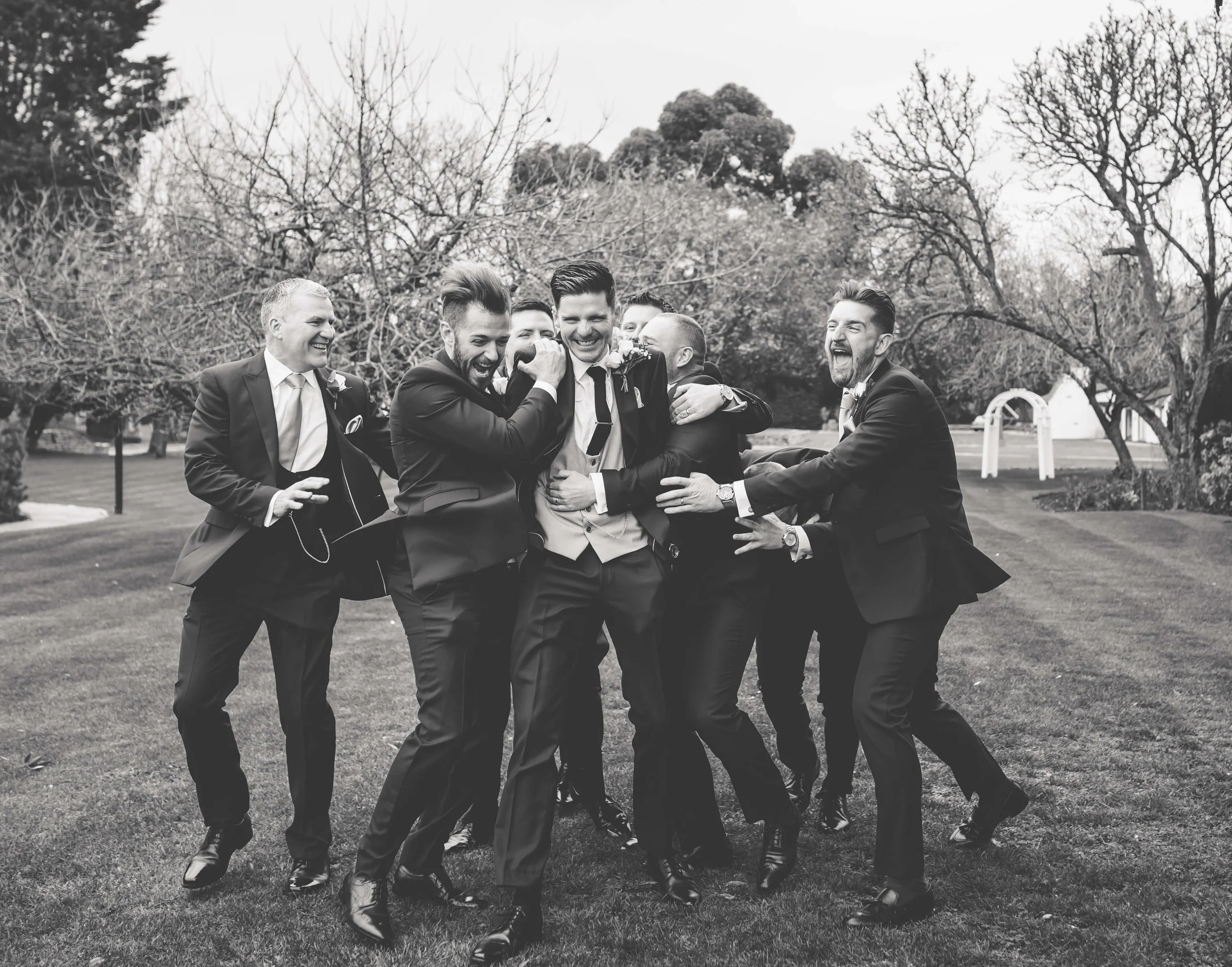 A group of men in suits celebrating and laughing outdoors on a lawn.