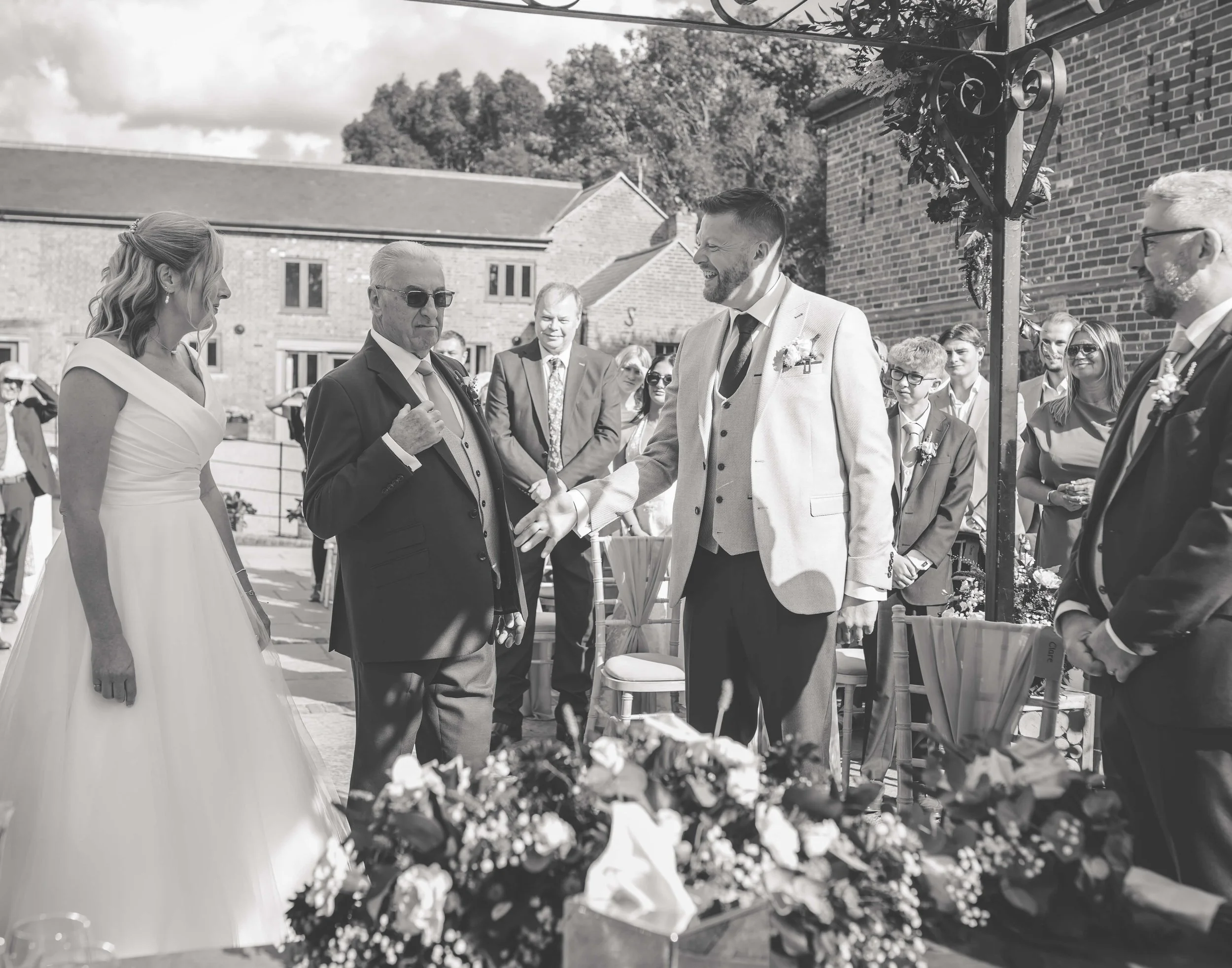 Black and white photo of a wedding ceremony outdoors with a bride in a white gown and a groom in a light-colored suit shaking hands with an older man in a suit, surrounded by guests.