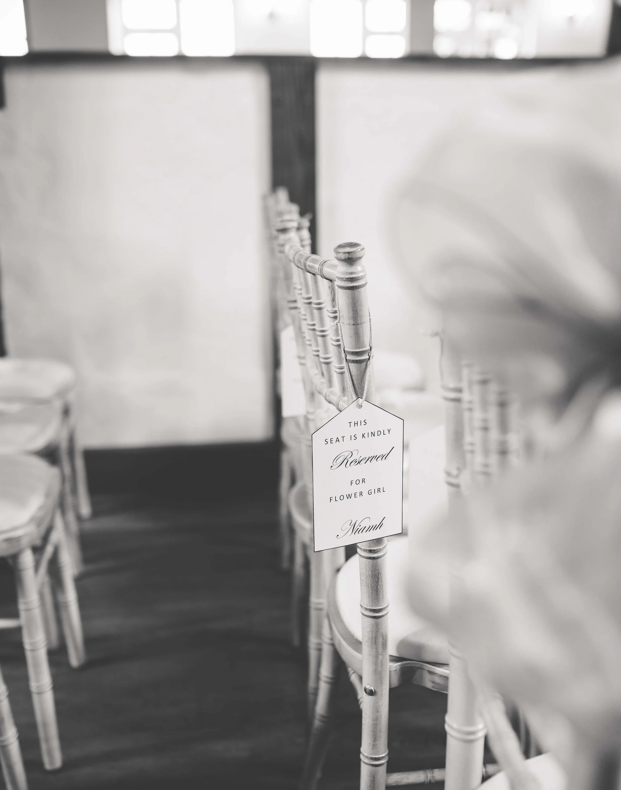 A row of white chairs decorated for a wedding, with a reserved sign reading "This seat is kindly reserved for Flower Girl Niamh."