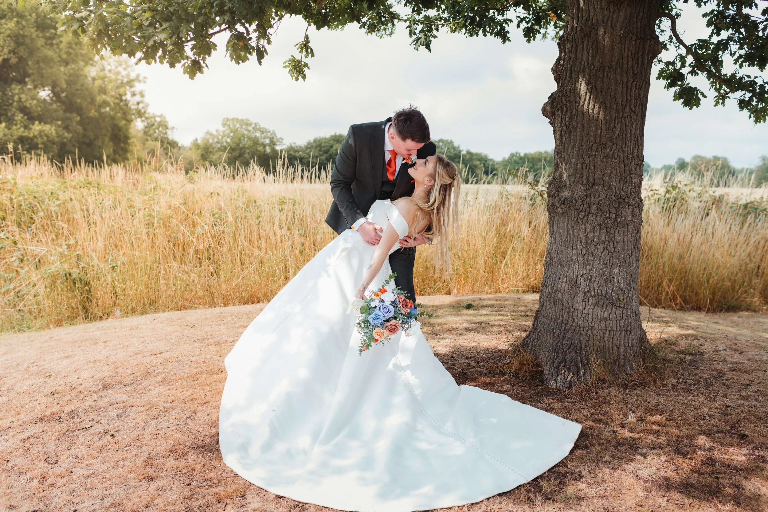 A bride and groom share a romantic moment outdoors, with the groom dipping the bride as they kiss under a large tree, surrounded by grassy fields.