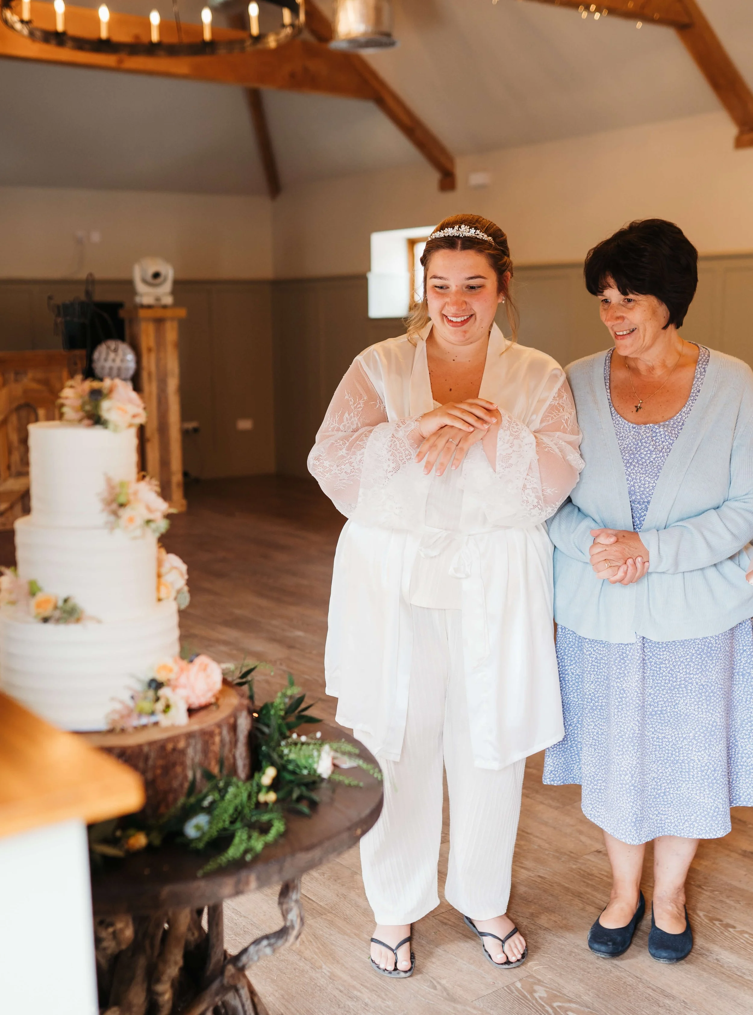 A woman in a white satin robe with lace sleeves and a crown of flowers on her head, smiling, standing next to an older woman in a light blue cardigan and a patterned blue dress, both looking at a wedding cake, in a rustic indoor setting.