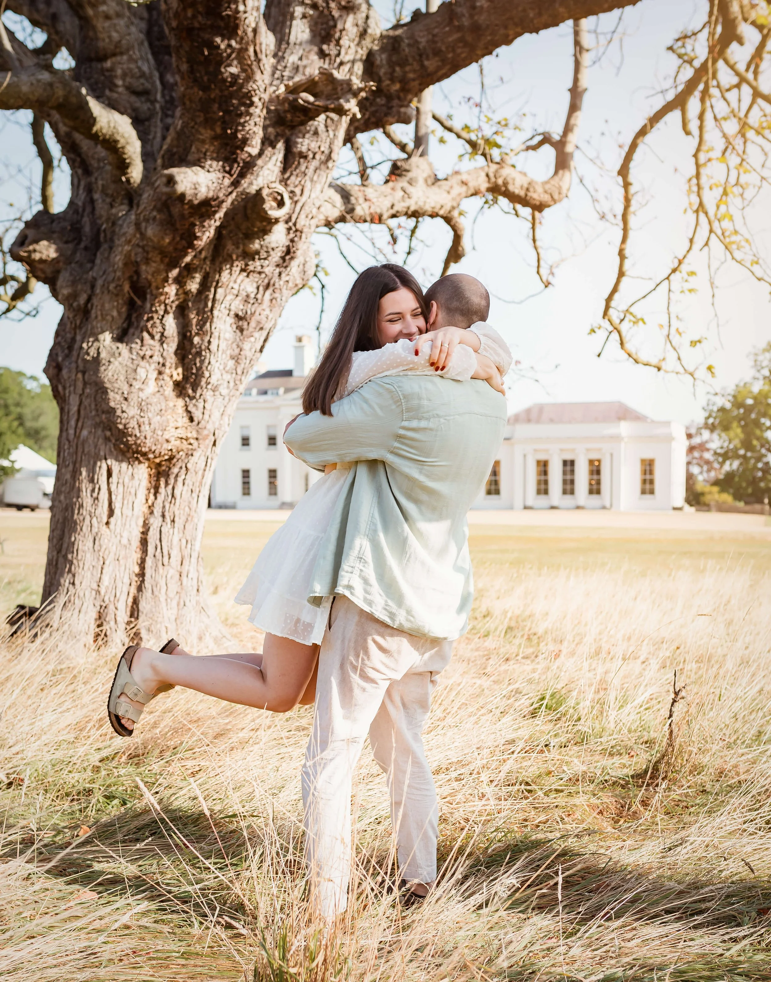 A man is lifting a woman in a white dress in a grassy field with a large tree nearby. They are hugging and smiling, with a white building in the background during what appears to be a sunny day.