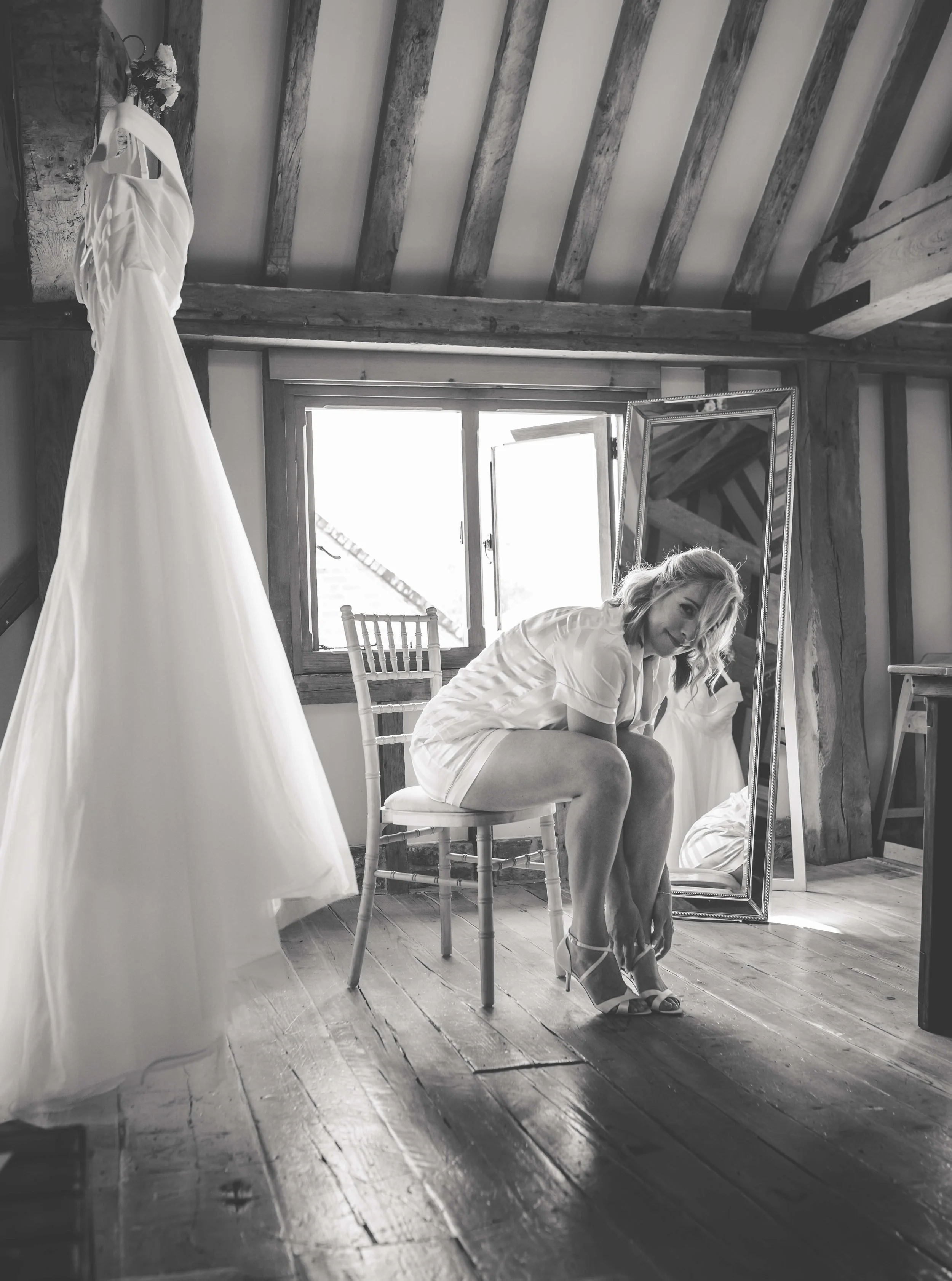 A woman sitting on a chair in a rustic room, trying on high-heeled shoes, with a wedding dress hanging nearby and a large mirror reflecting her image.