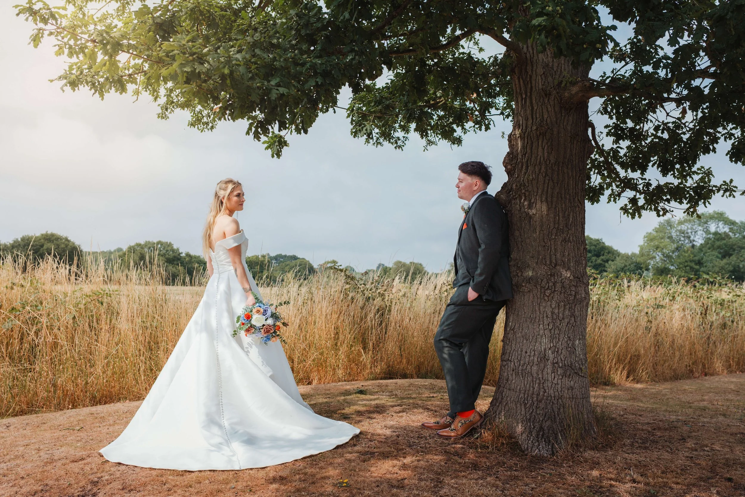 bride and groom look on lovingly at each other as groom leans casually against the tree 