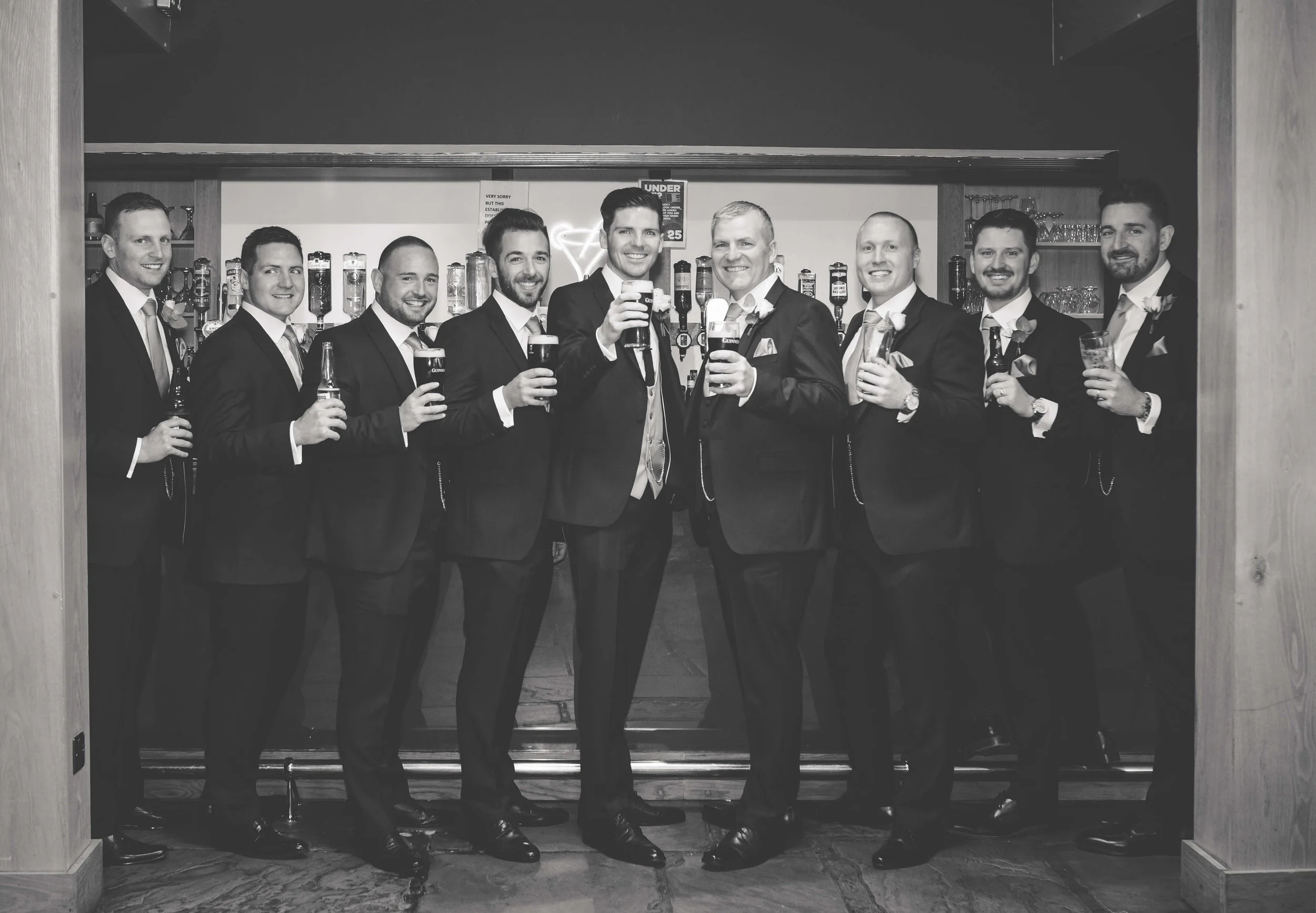 A group of nine men dressed in suits celebrating with drinks in a bar or pub.