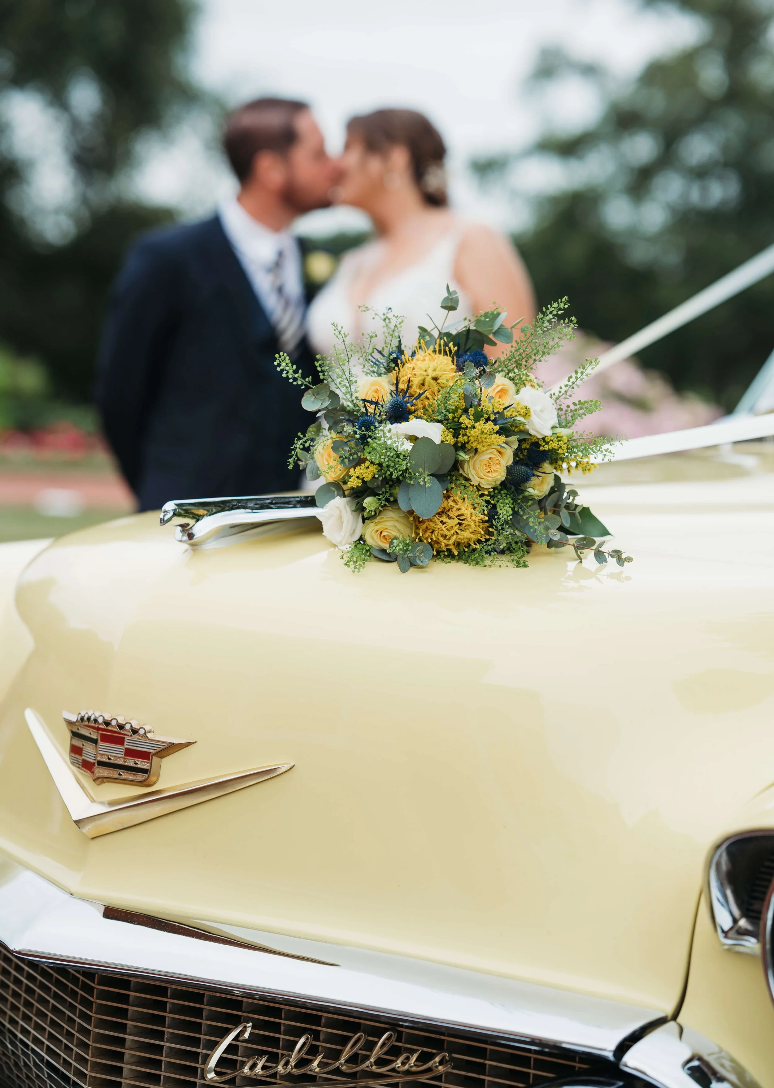 A yellow Cadillac car with a flower arrangement on the hood, featuring yellow roses, white flowers, greenery, and dark blue accents. In the background, a bride and groom in wedding attire are kissing, out of focus.