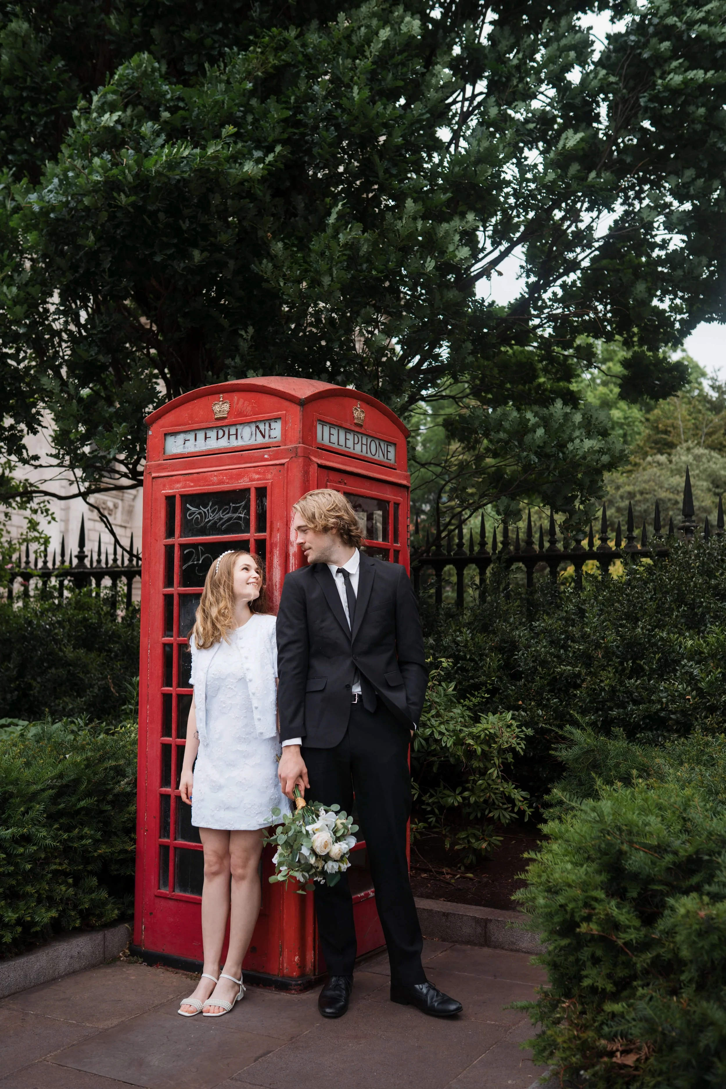 A young woman and man in formal attire standing beside a red British telephone booth in a park with lush green trees and shrubs, and a black wrought iron fence in the background. The woman is in a white dress holding a bouquet of flowers, and the man