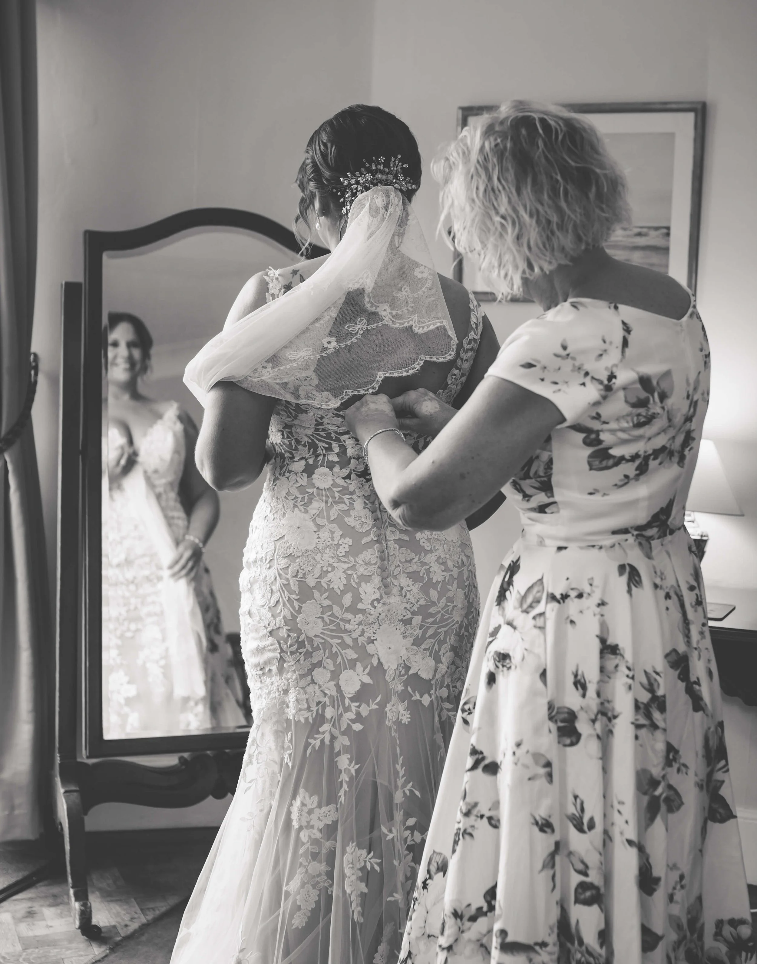 A bride helps a woman fasten her wedding dress in front of a mirror.