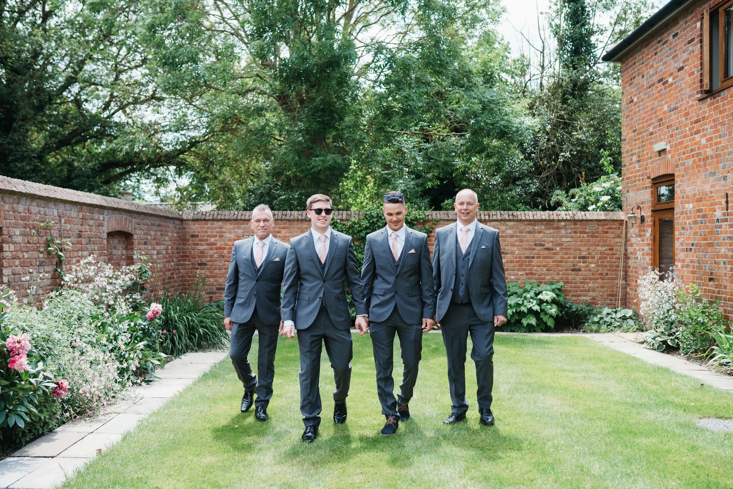 Group of four men dressed in gray suits, white shirts, and pink ties walking on a lawn in a garden with brick walls and trees in the background.