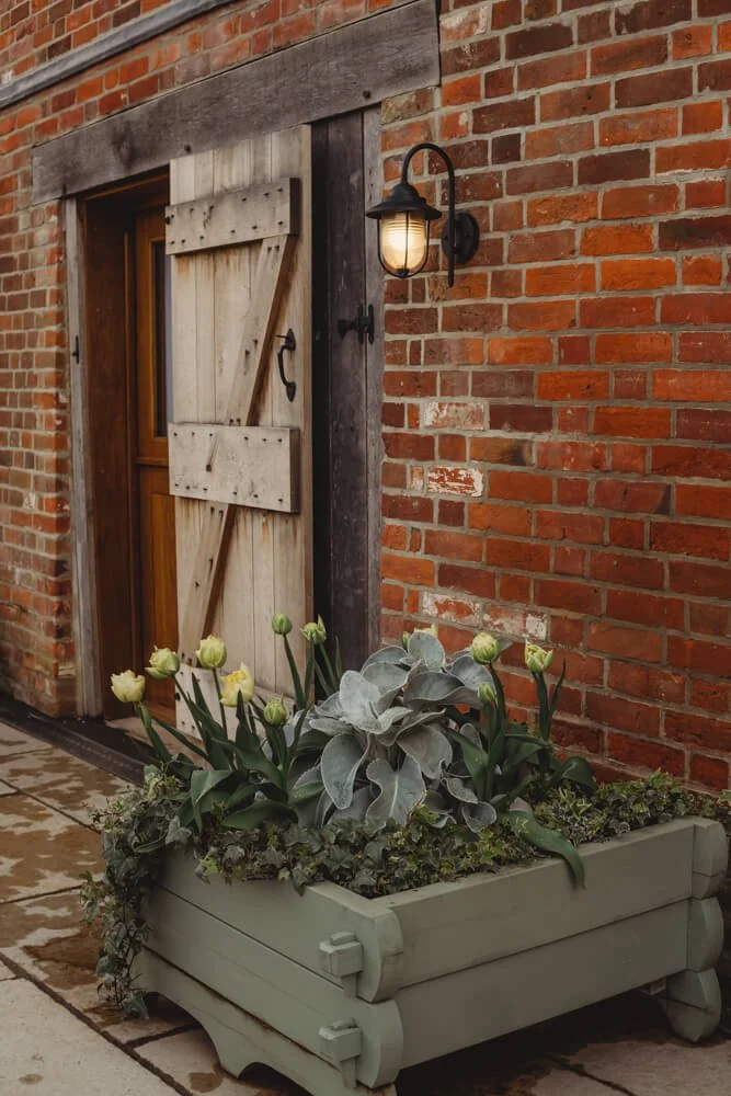 A rustic wooden door with black hardware on a brick wall exterior, illuminated by a black outdoor wall lantern. In front, there is a large decorative planter box filled with green plants and white tulips, placed on a stone sidewalk.