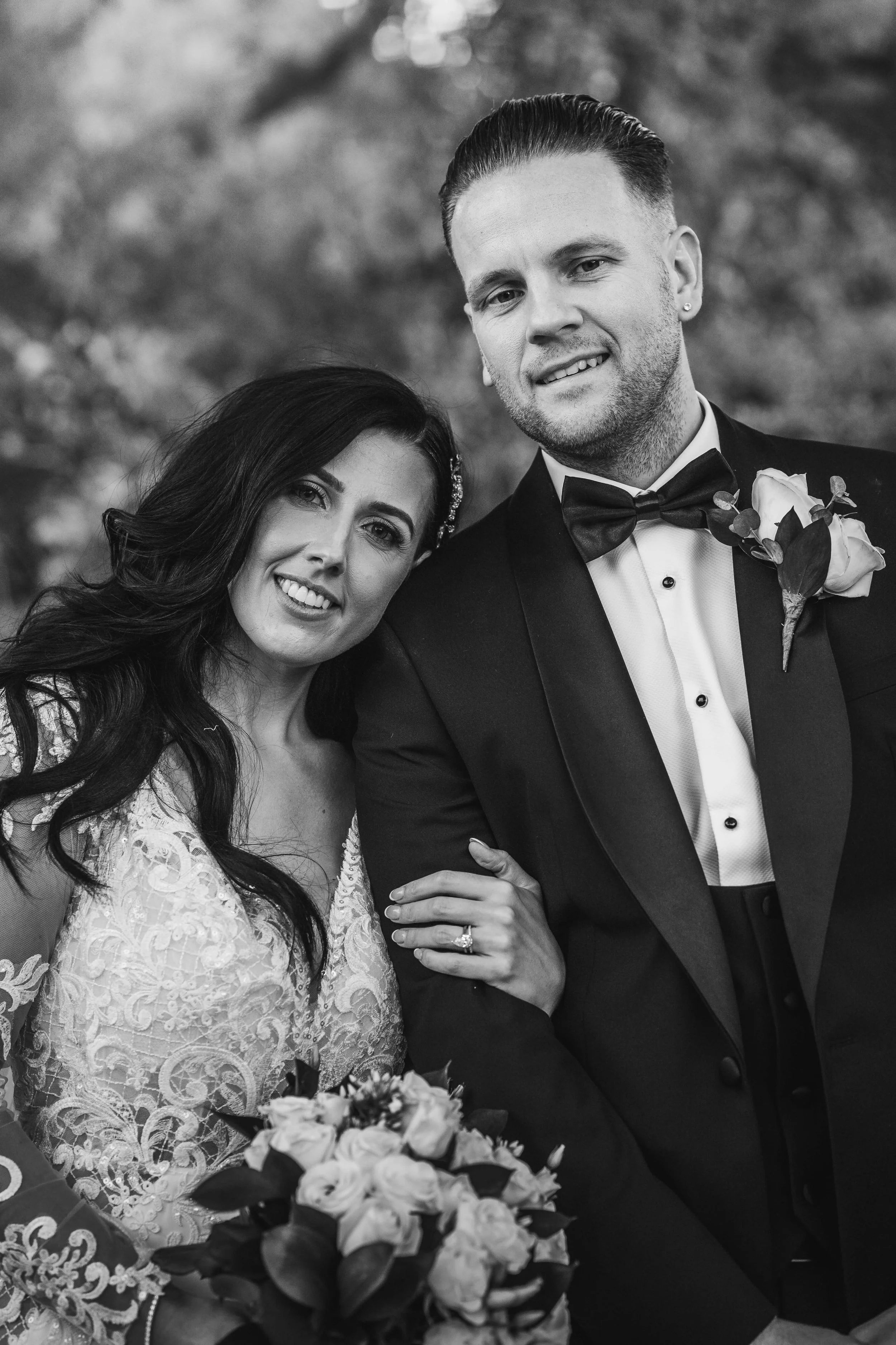 Black and white wedding photo of a bride and groom outdoors. The bride has long dark hair and is wearing a lace wedding dress, holding a bouquet of roses. The groom has short hair, a beard, and is wearing a tuxedo with a bow tie and a boutonniere. Th