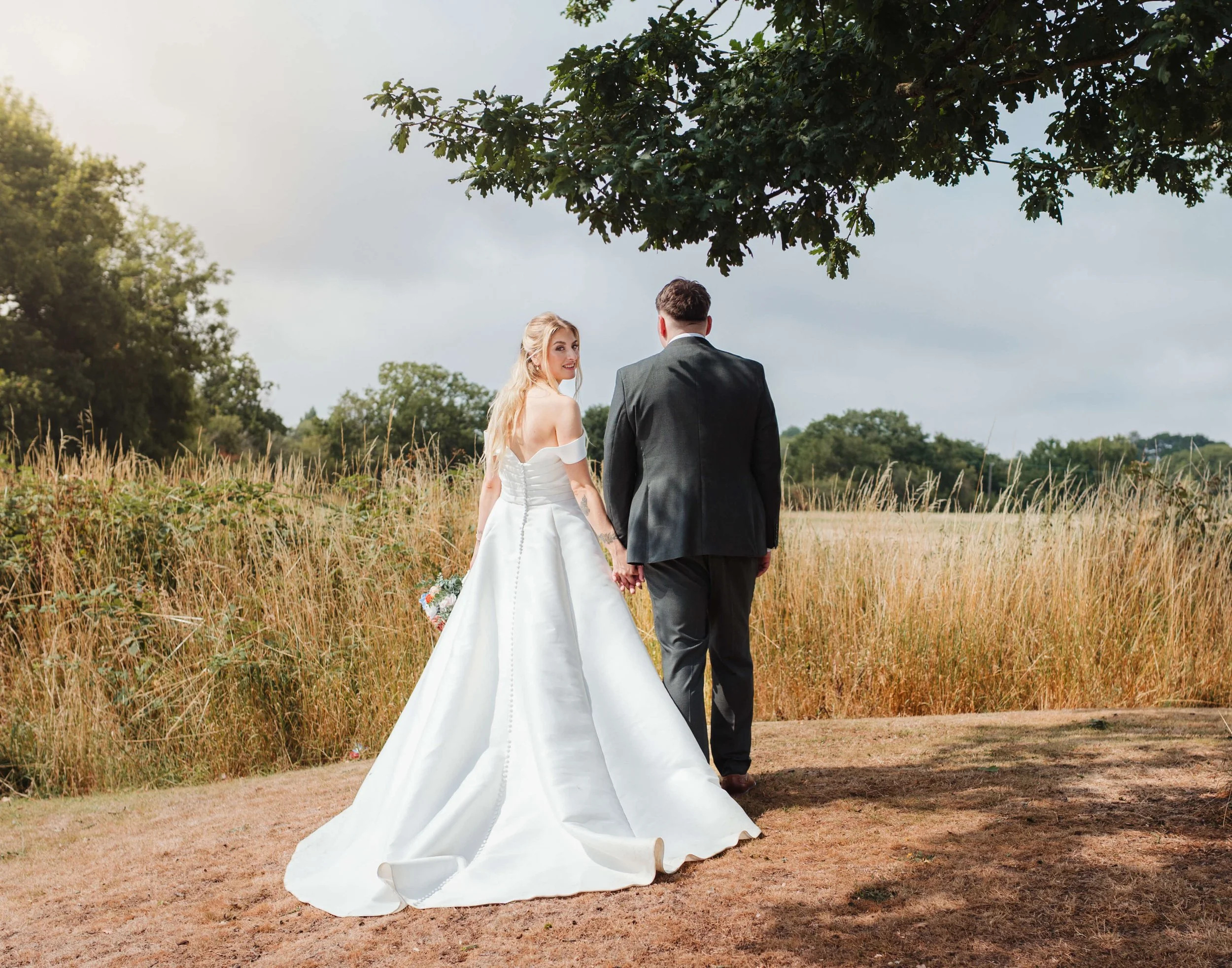 A bride and groom holding hands in an open field during their wedding day, with trees and a cloudy sky in the background.