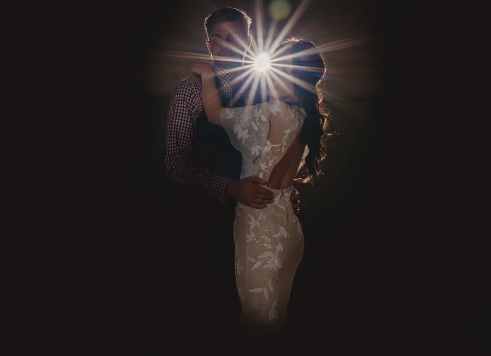 A couple is dancing at a wedding, with a bright light shining behind them, obscuring their faces.