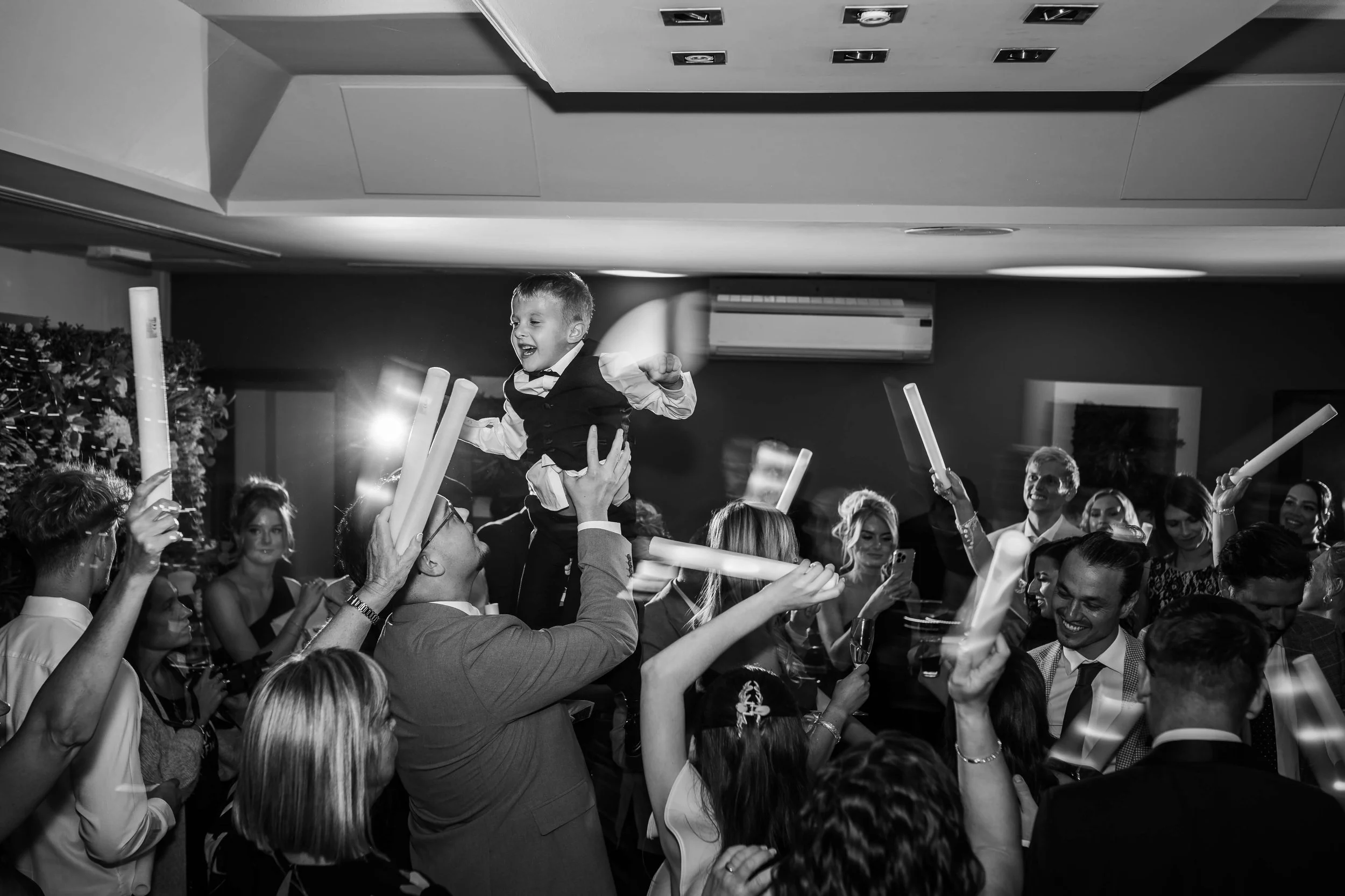 Celebration scene with a young boy being lifted in a joyful crowd, everyone holding foam sticks at a party or event, in black and white.