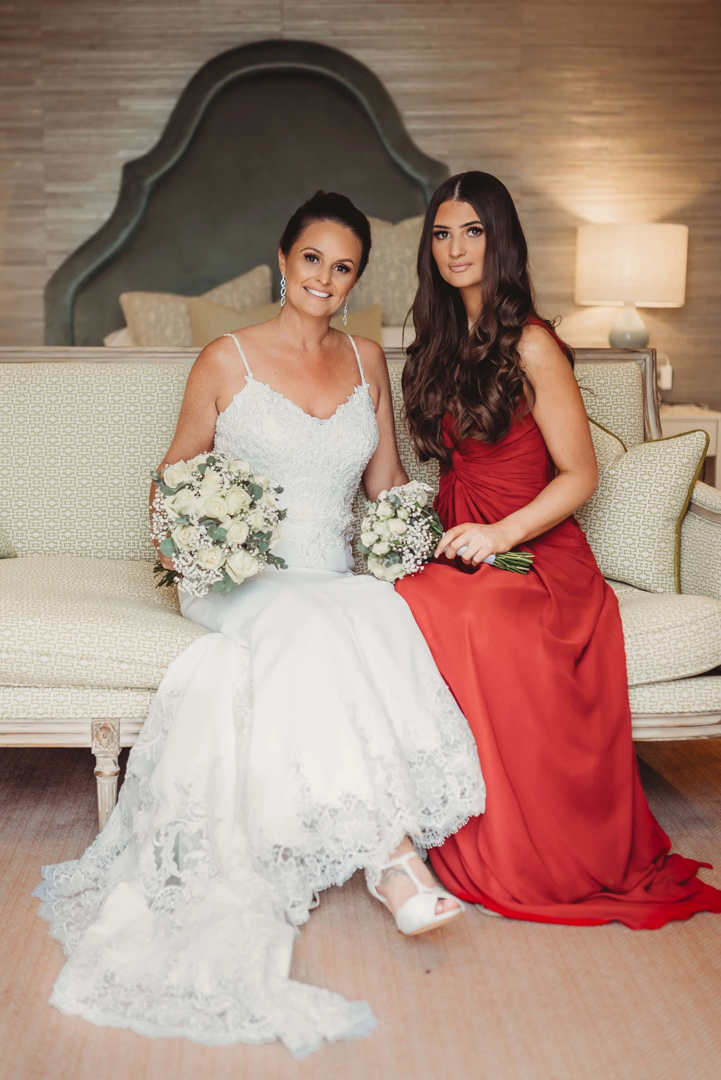 A bride in a white lace wedding dress sitting on a sofa next to a woman in a red gown, both holding bouquets of white roses and greenery, indoors with beige and brown decor.