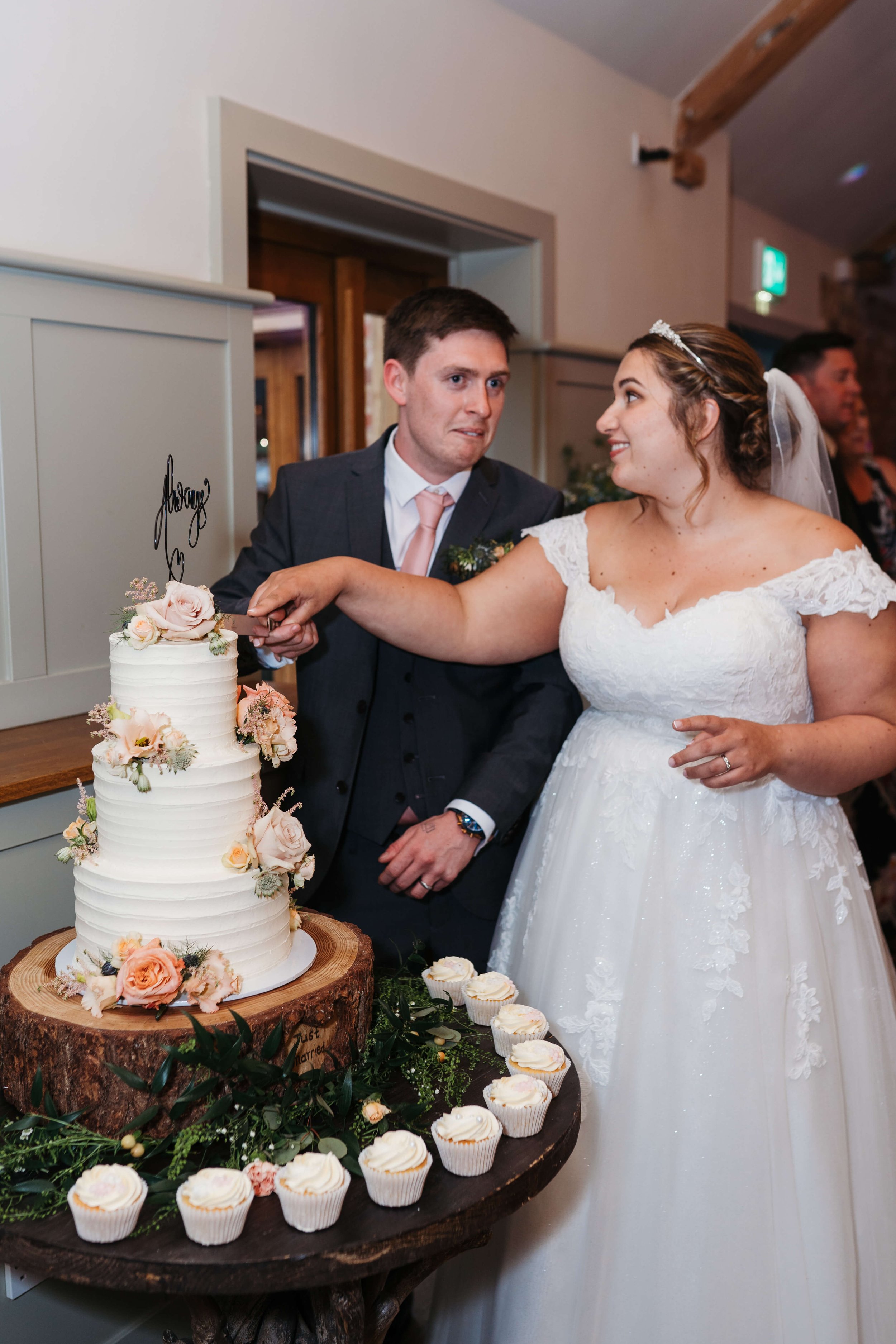 A bride and groom cutting a wedding cake together, with wedding cupcakes on a rustic wooden table in front of them, at their wedding reception in a cozy indoor venue.