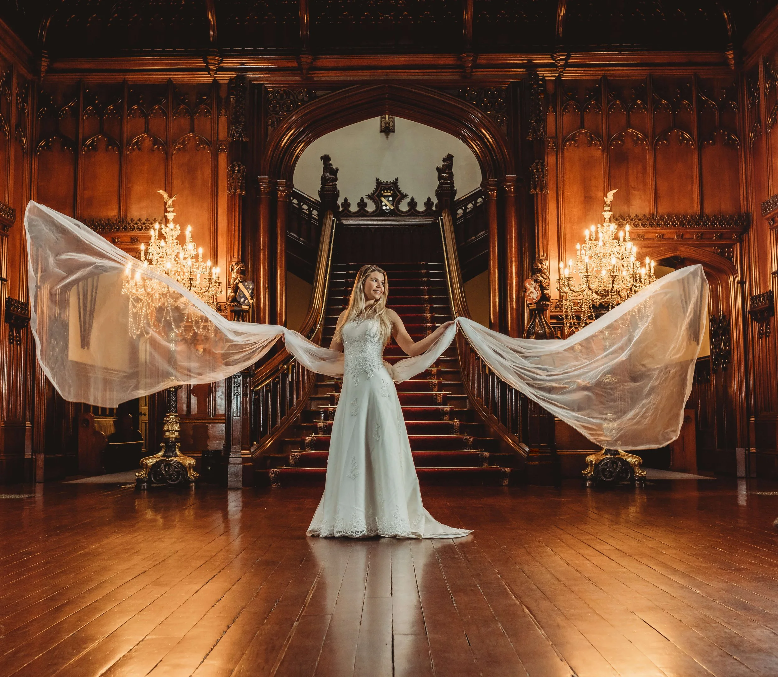 A woman in a white wedding dress stands on a grand wooden staircase inside a lavishly decorated room with ornate wood paneling, chandeliers, and a polished wooden floor. She is holding a sheer, flowing veil out to her sides, smiling and looking to he