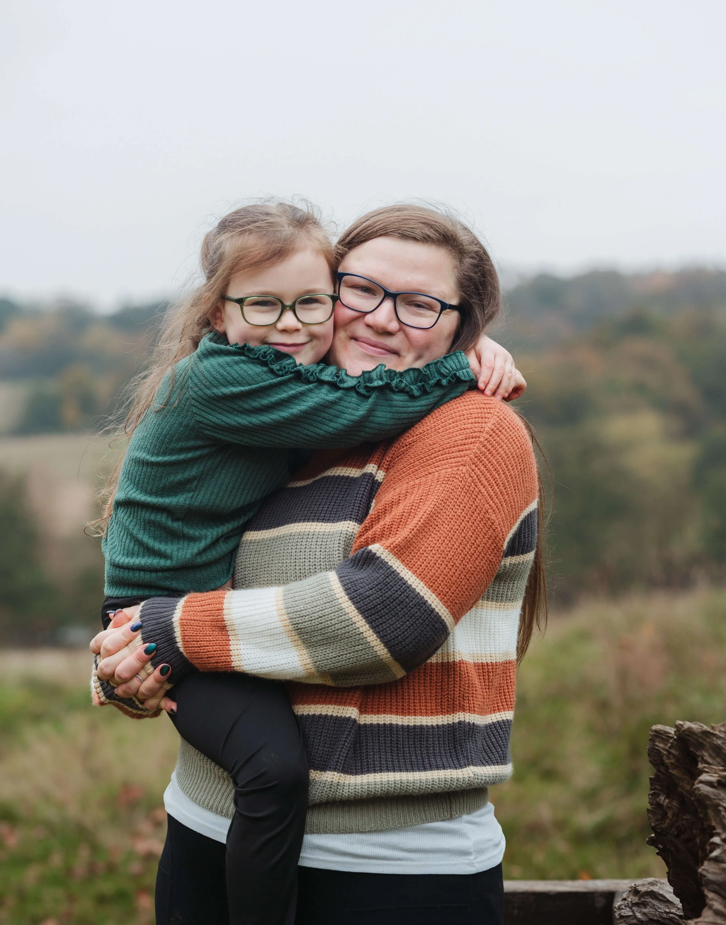 A woman in glasses holding a young girl, both smiling and hugging outdoors in a rural setting.
