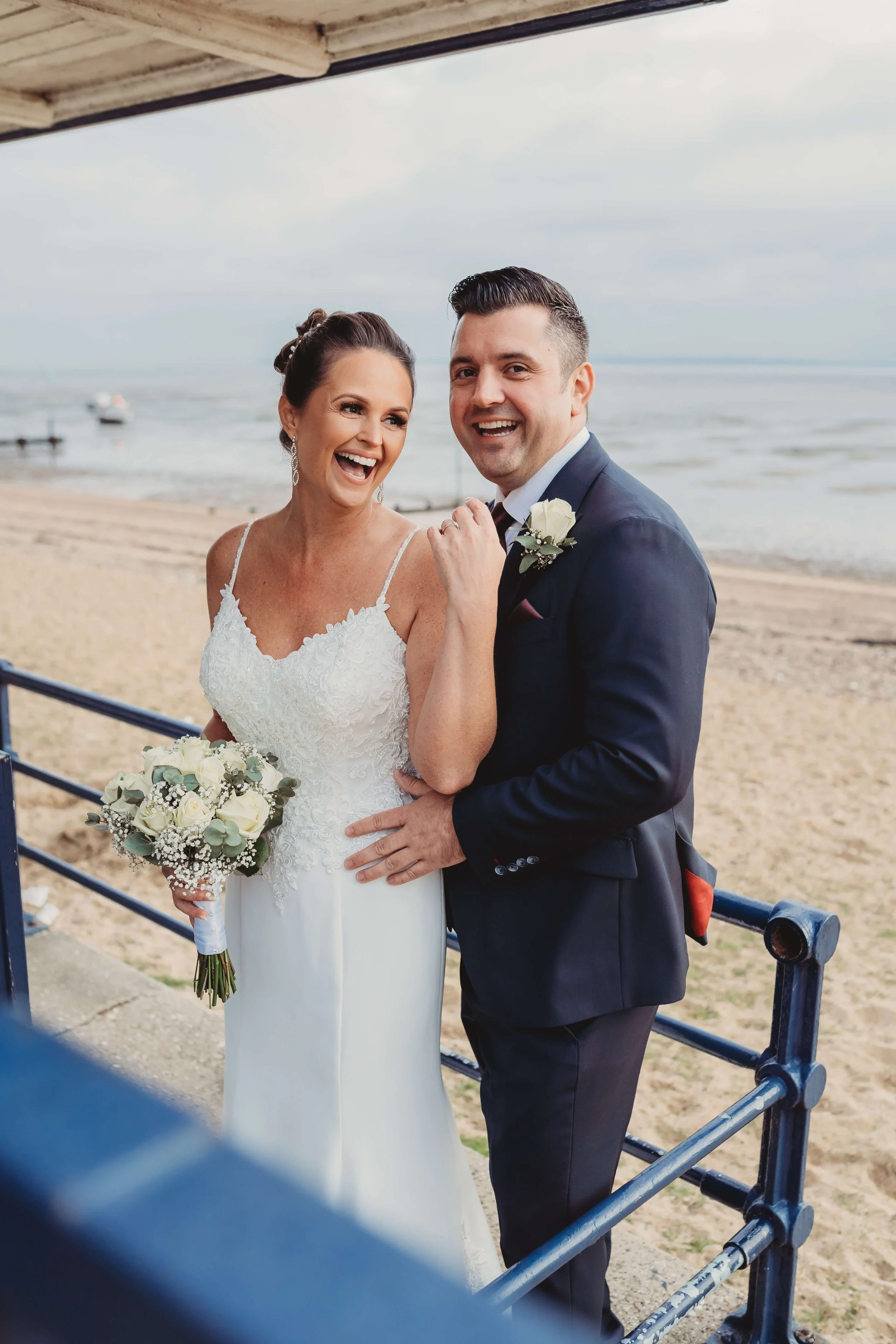 A bride and groom celebrating their wedding by the beach, smiling happily and holding each other.