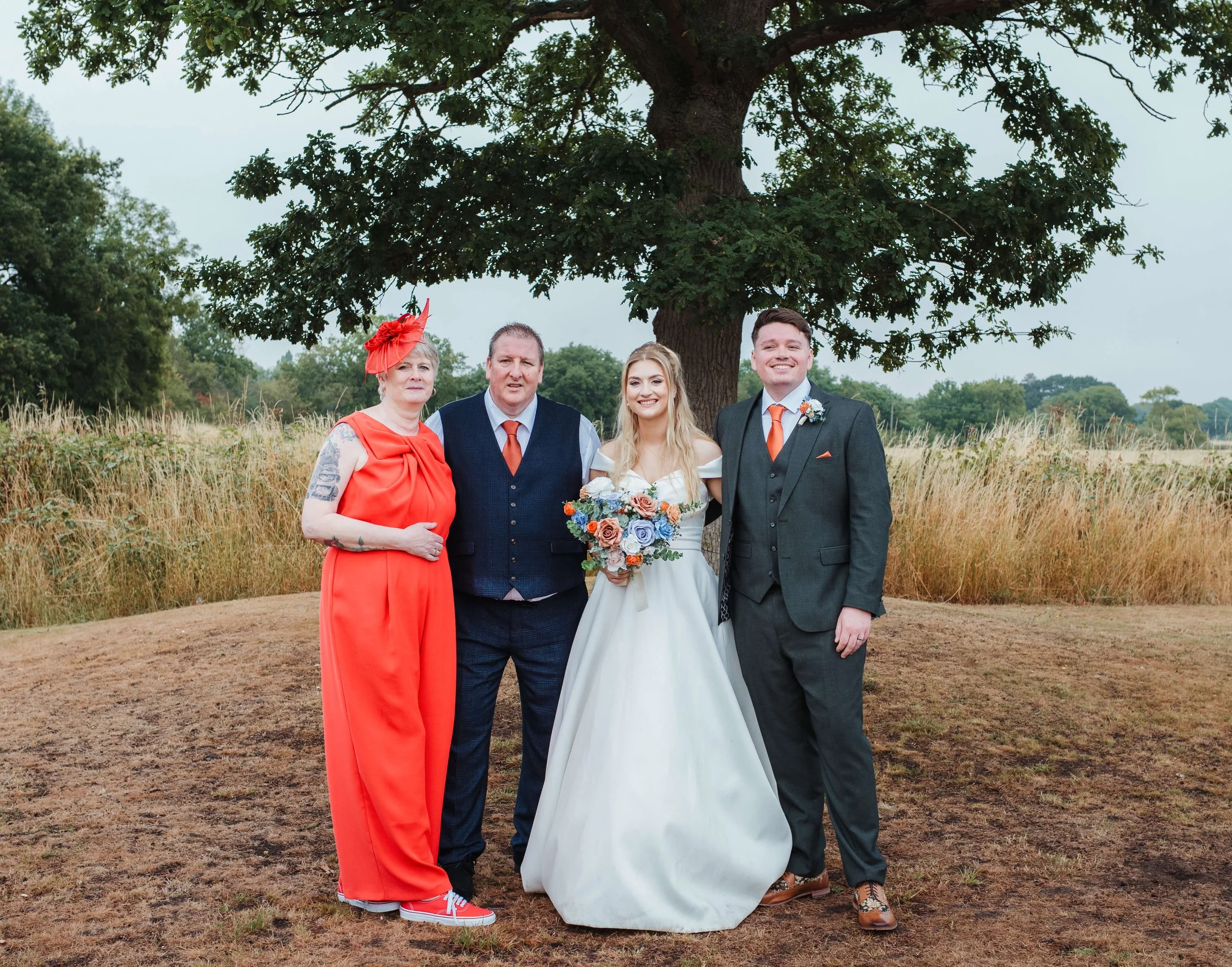 A group of four people standing outdoors beneath a large tree, with a field and tall grass in the background, at a wedding celebration. The bride, wearing a white wedding dress and holding a bouquet of colorful flowers, stands next to the groom, who 