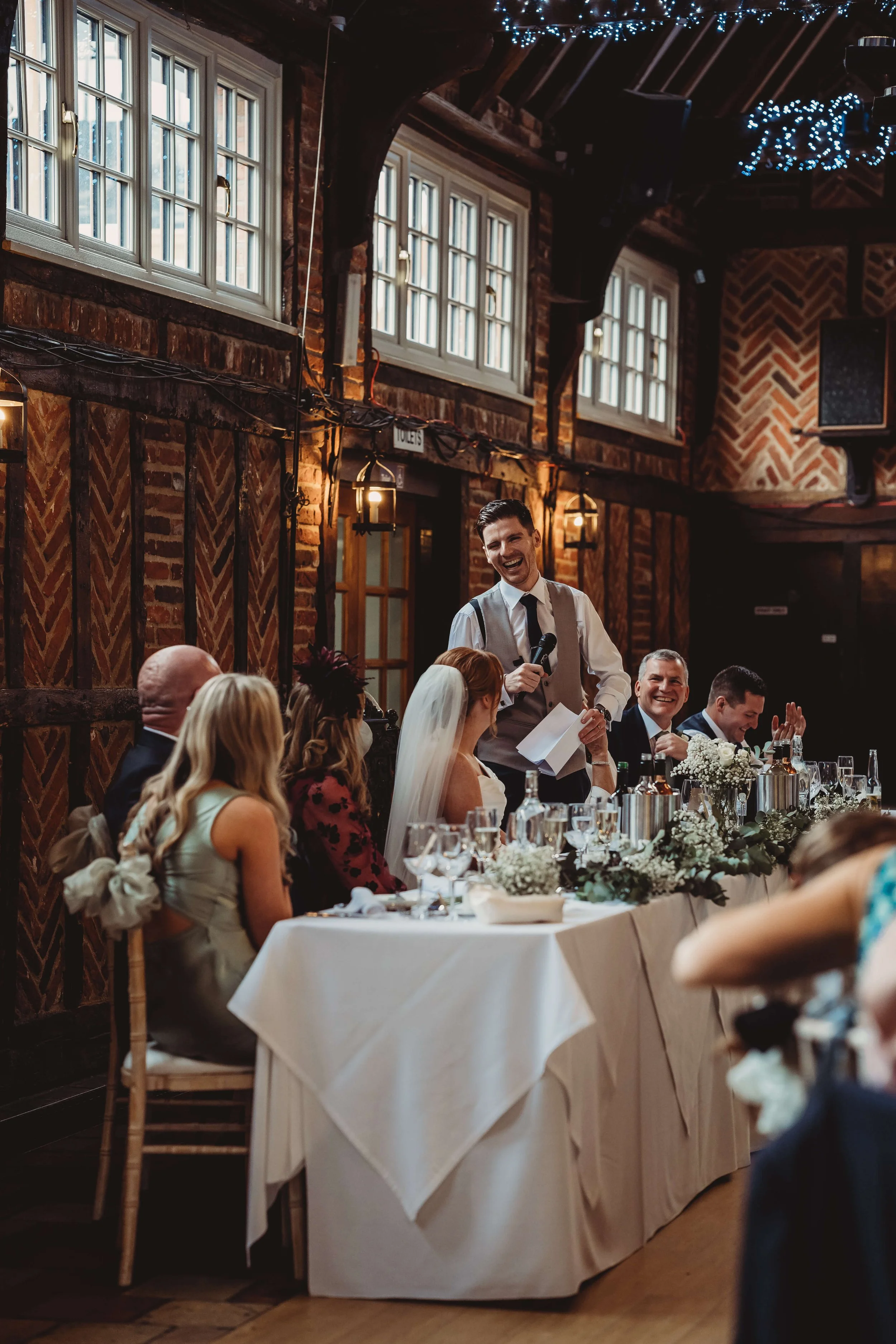 A man giving a speech at a wedding reception table with bridesmaids and groomsmen seated, decorated with flowers, in a rustic indoor venue with brick walls and large windows.