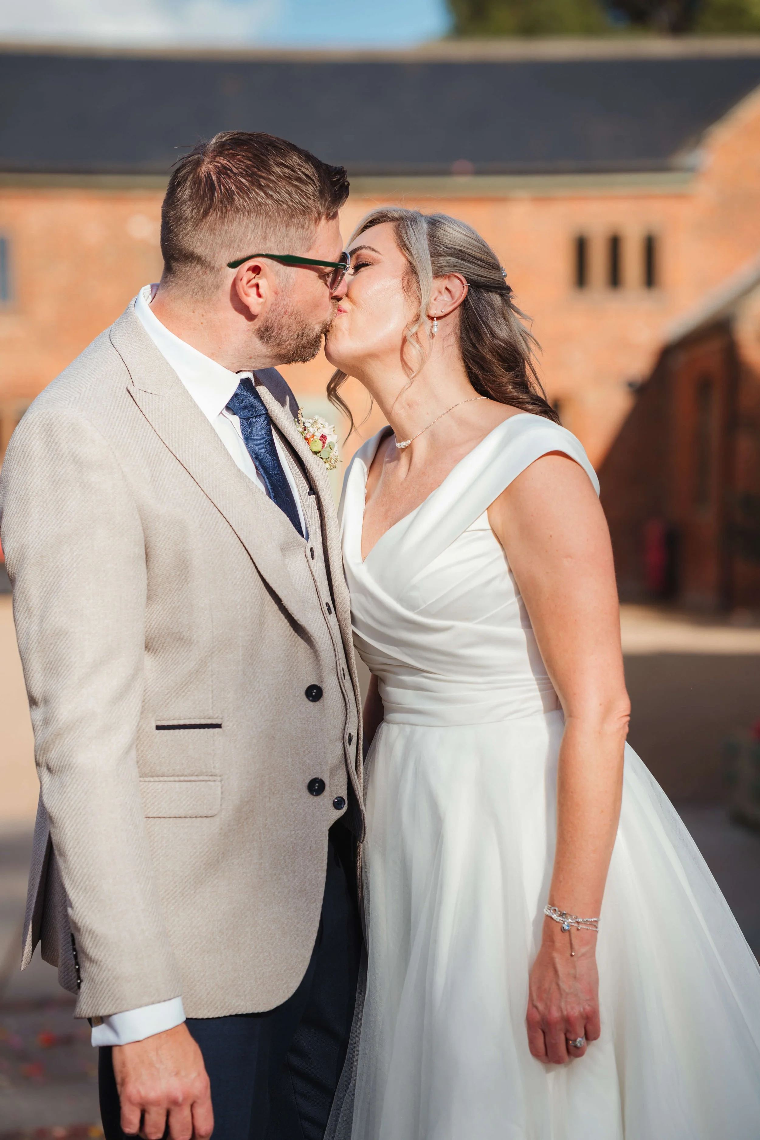 A bride and groom kiss outdoors in front of a brick building on their wedding day.