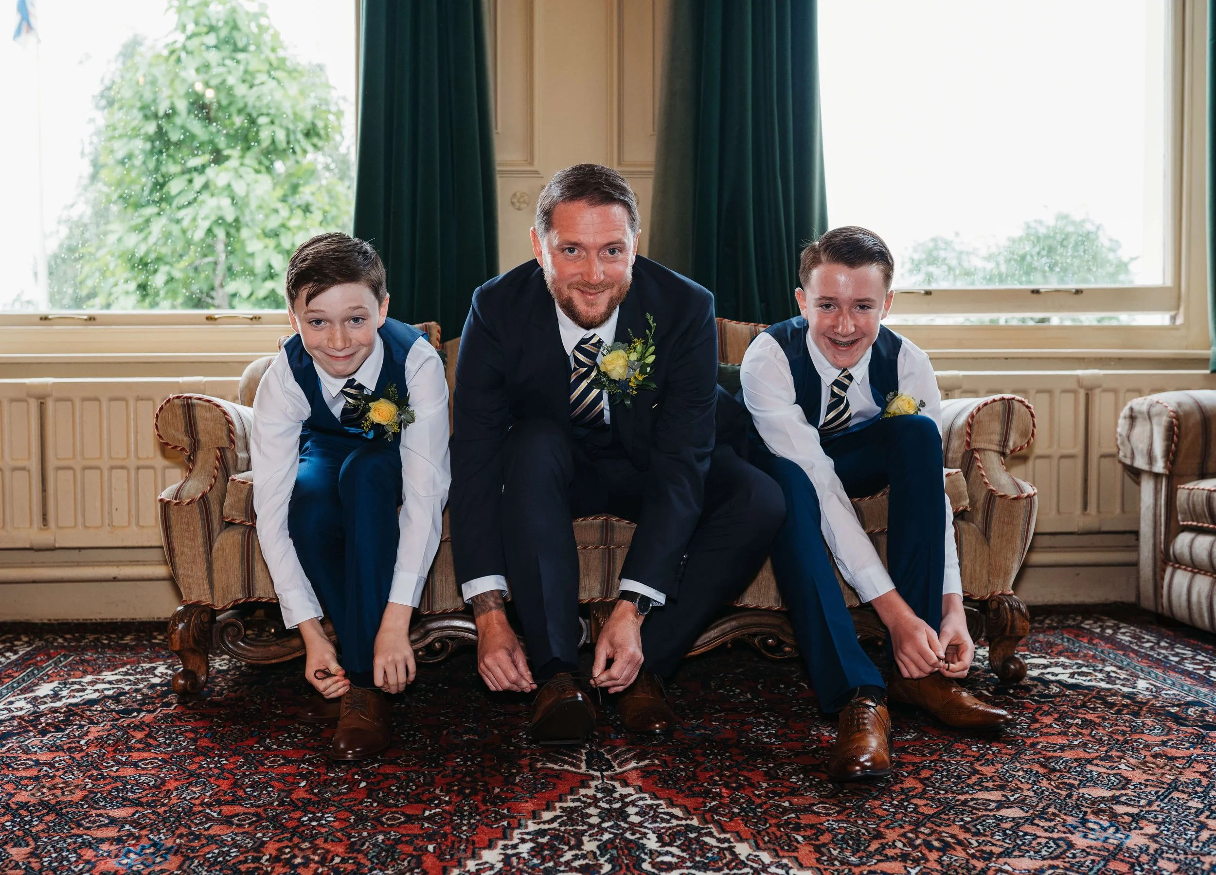 A man and two boys, dressed in formal attire with matching striped ties and boutonnieres, are sitting on a vintage sofa indoors, tying their shoelaces. They are smiling and looking at the camera, with large windows and green curtains behind them, and