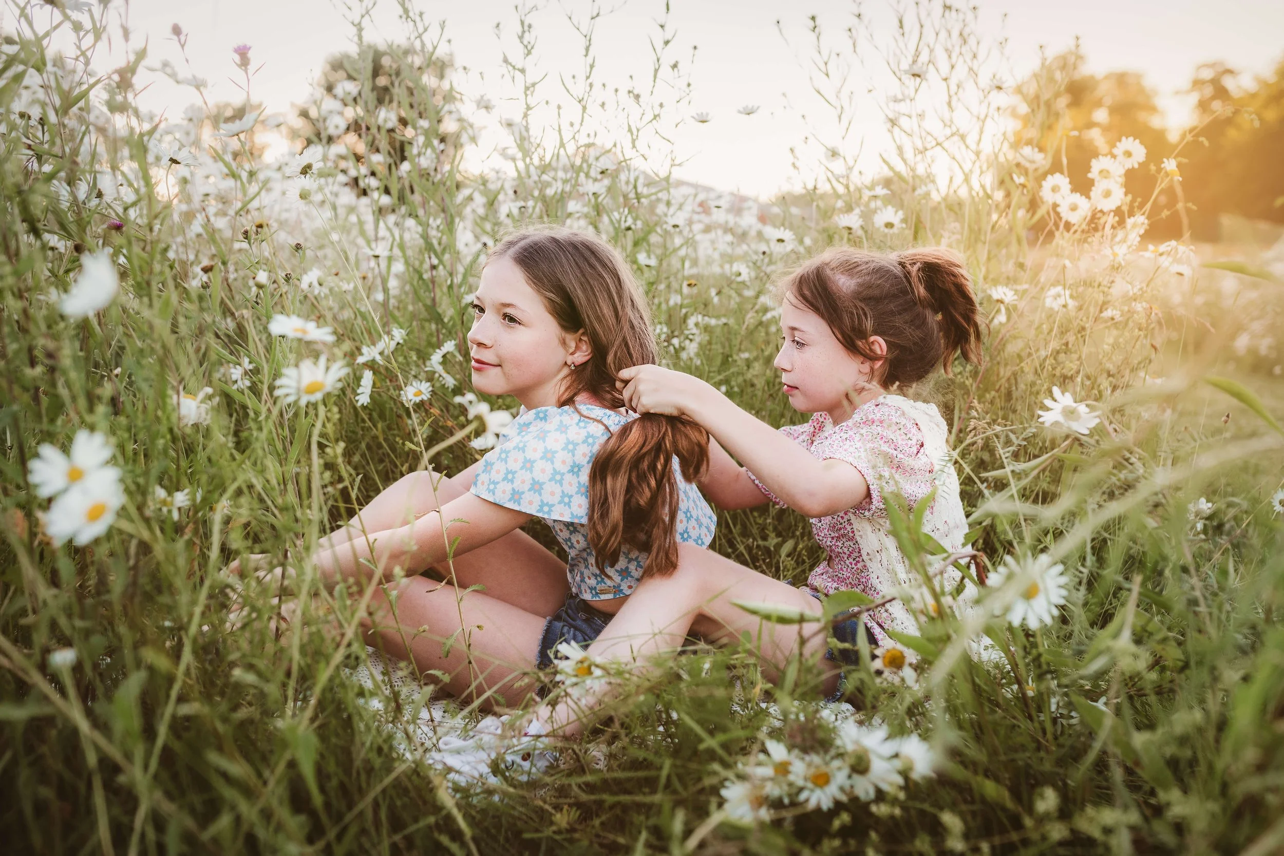 Two young girls sitting in a field of daisies, one girl braiding the other’s hair during sunset.