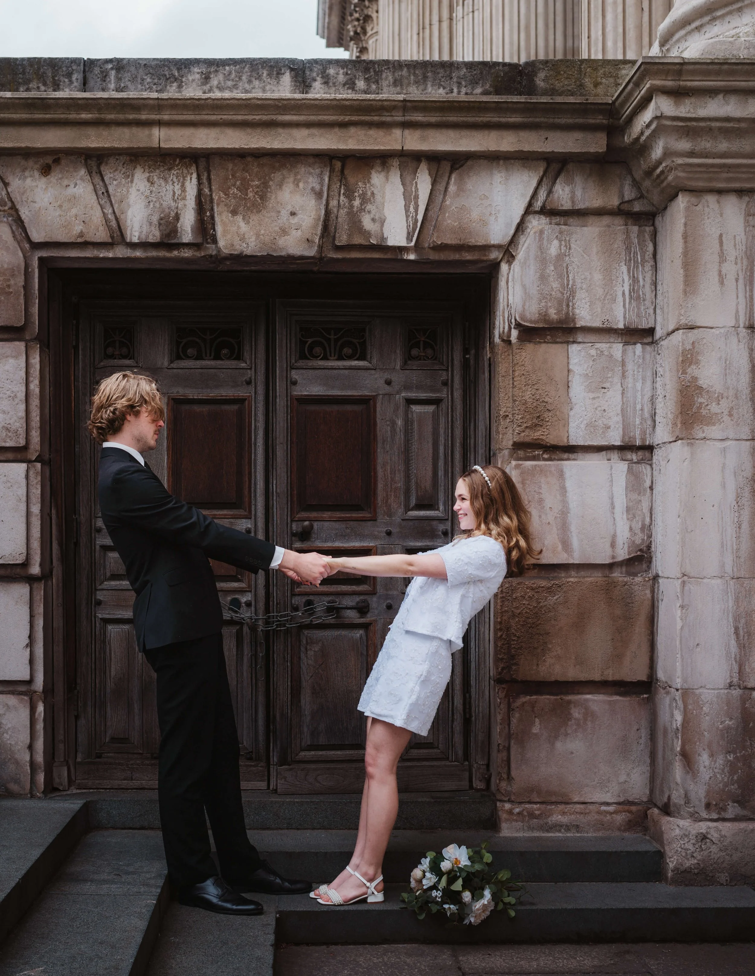 A couple dressed in wedding attire holding hands in front of a large wooden door, with a bouquet of flowers on the ground nearby.