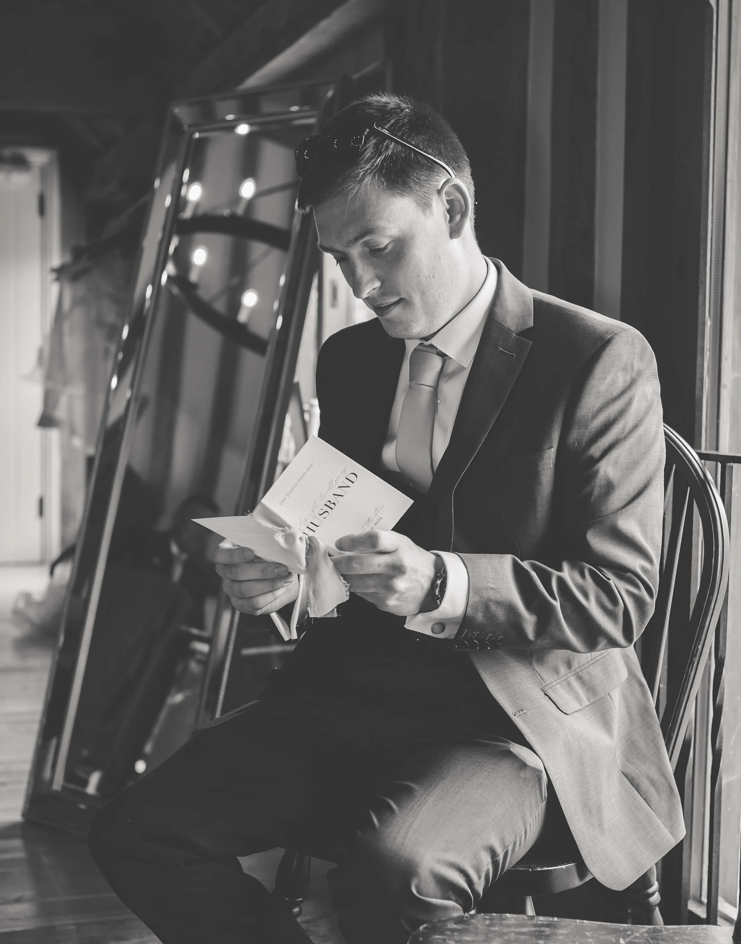 A man in a suit sitting on a chair, reading a wedding invitation card.