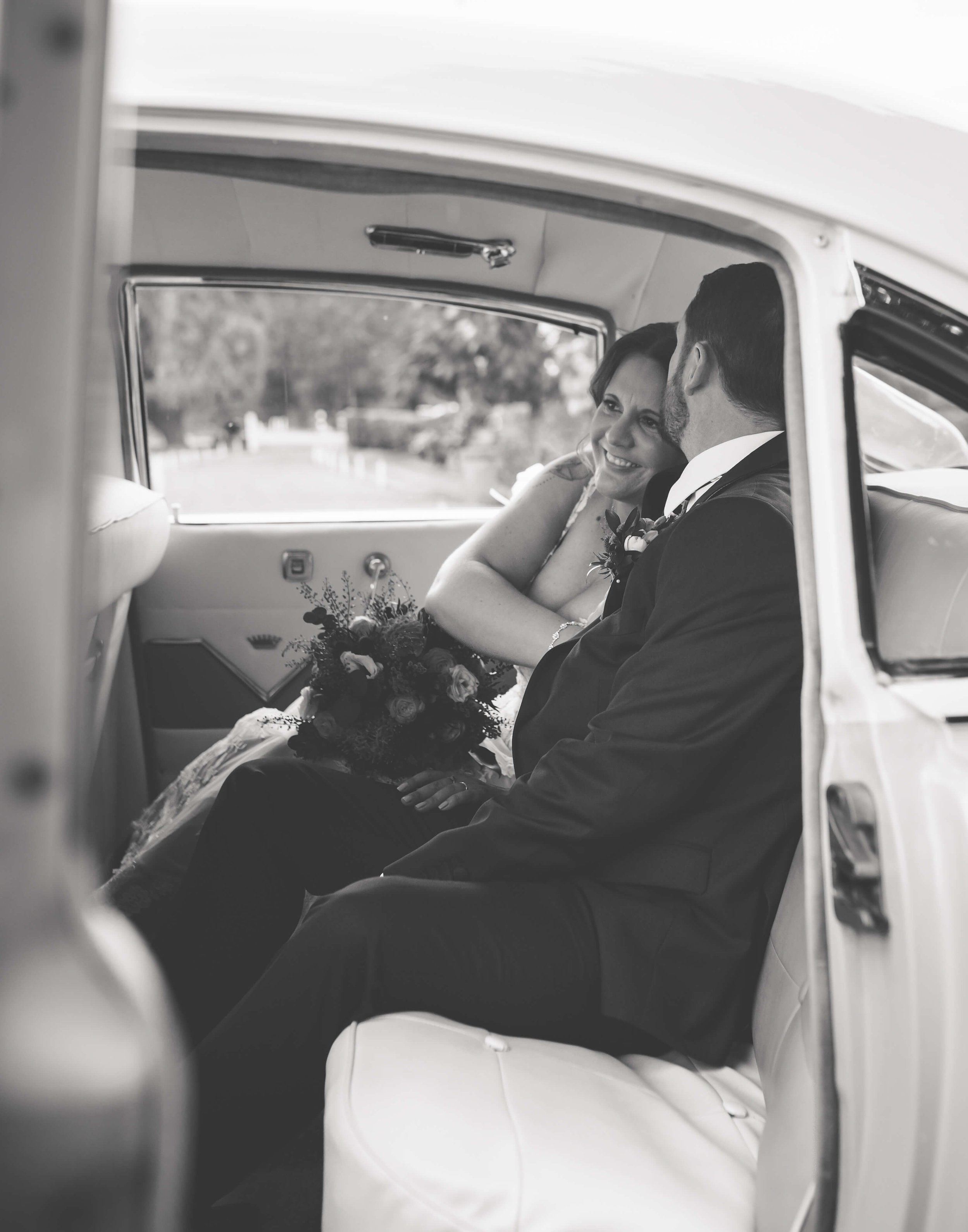 Black and white photo of a bride and groom sitting together inside a vintage car, smiling and looking at each other, with the bride holding a bouquet of flowers.
