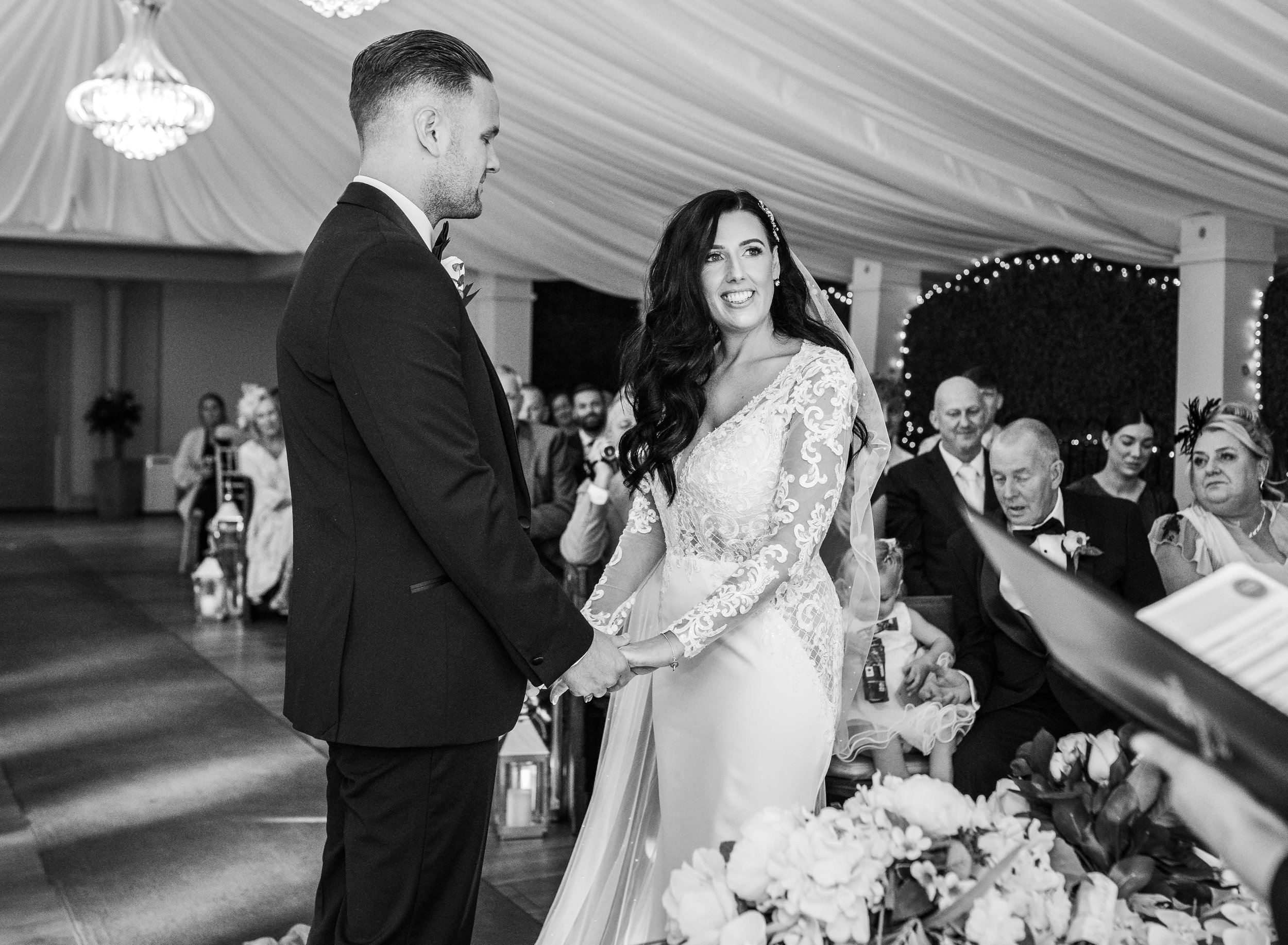 black and white photo of bride and groom at the head of the aisle joining hands 