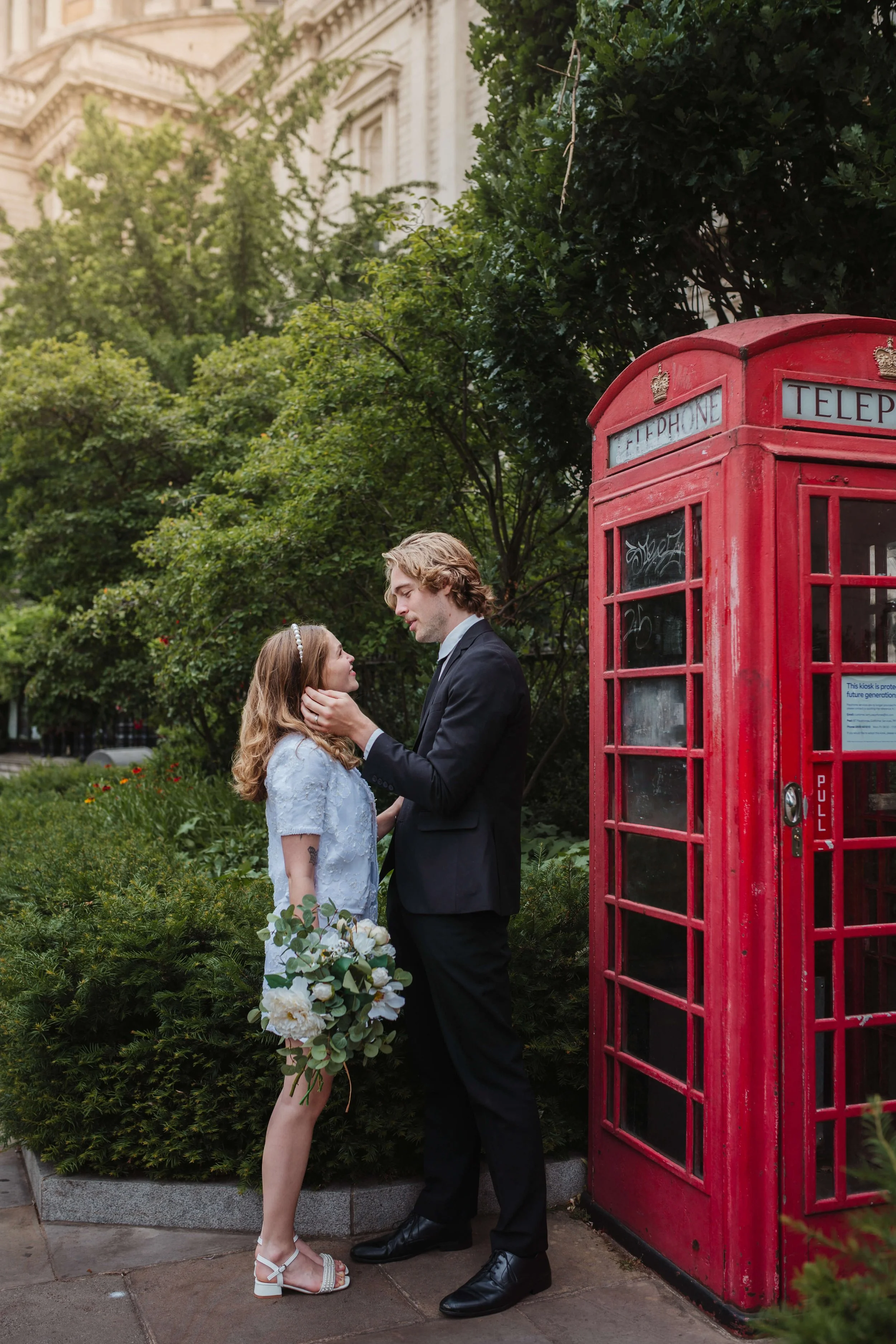 A young couple in wedding attire standing close to each other beside a red telephone booth in a lush green park, with the man gently holding the woman's face.
