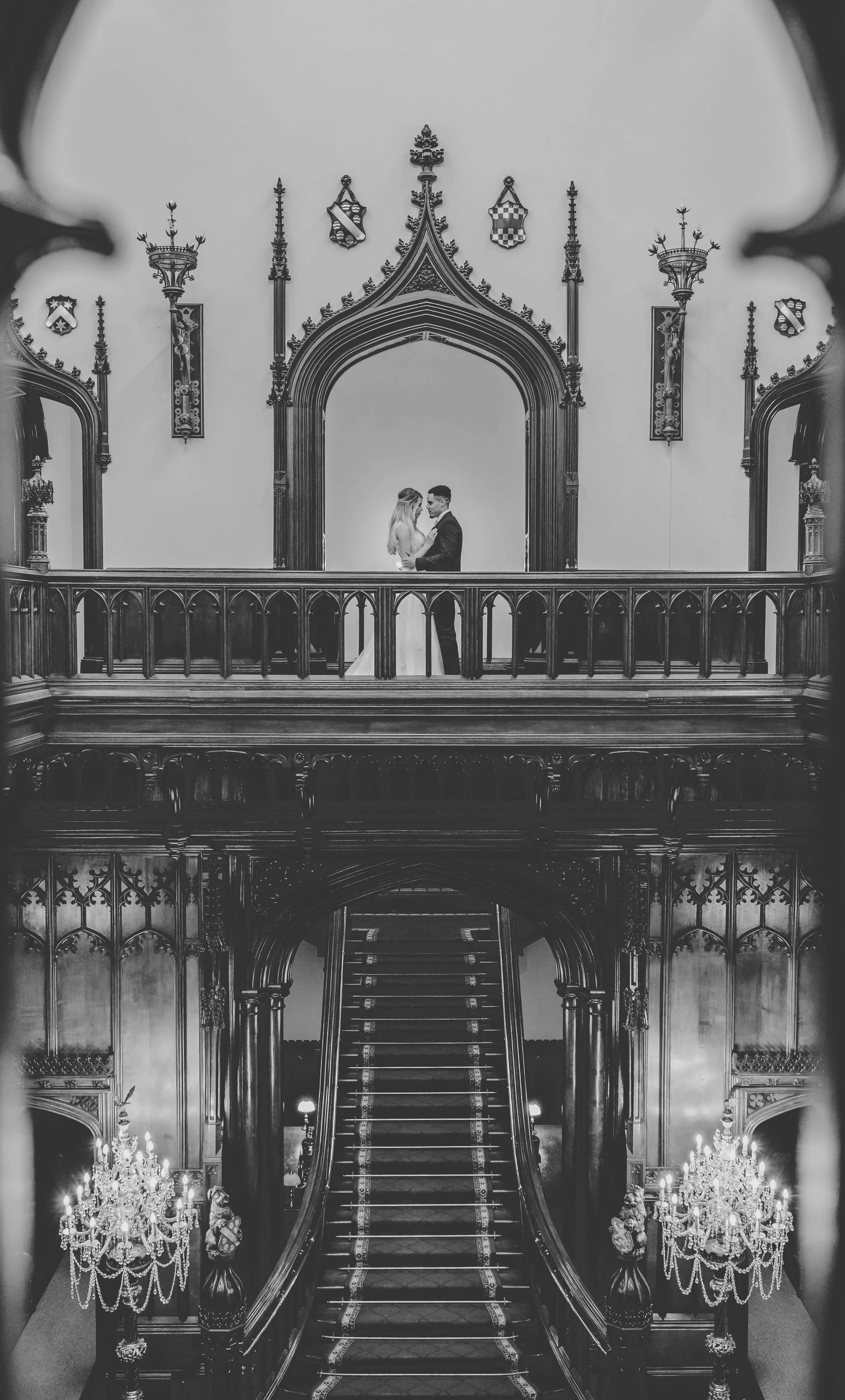 A black and white photo of a wedding couple standing on an ornate balcony inside a grand, gothic-style building with intricate woodwork, chandeliers, and decorative shields on the wall.
