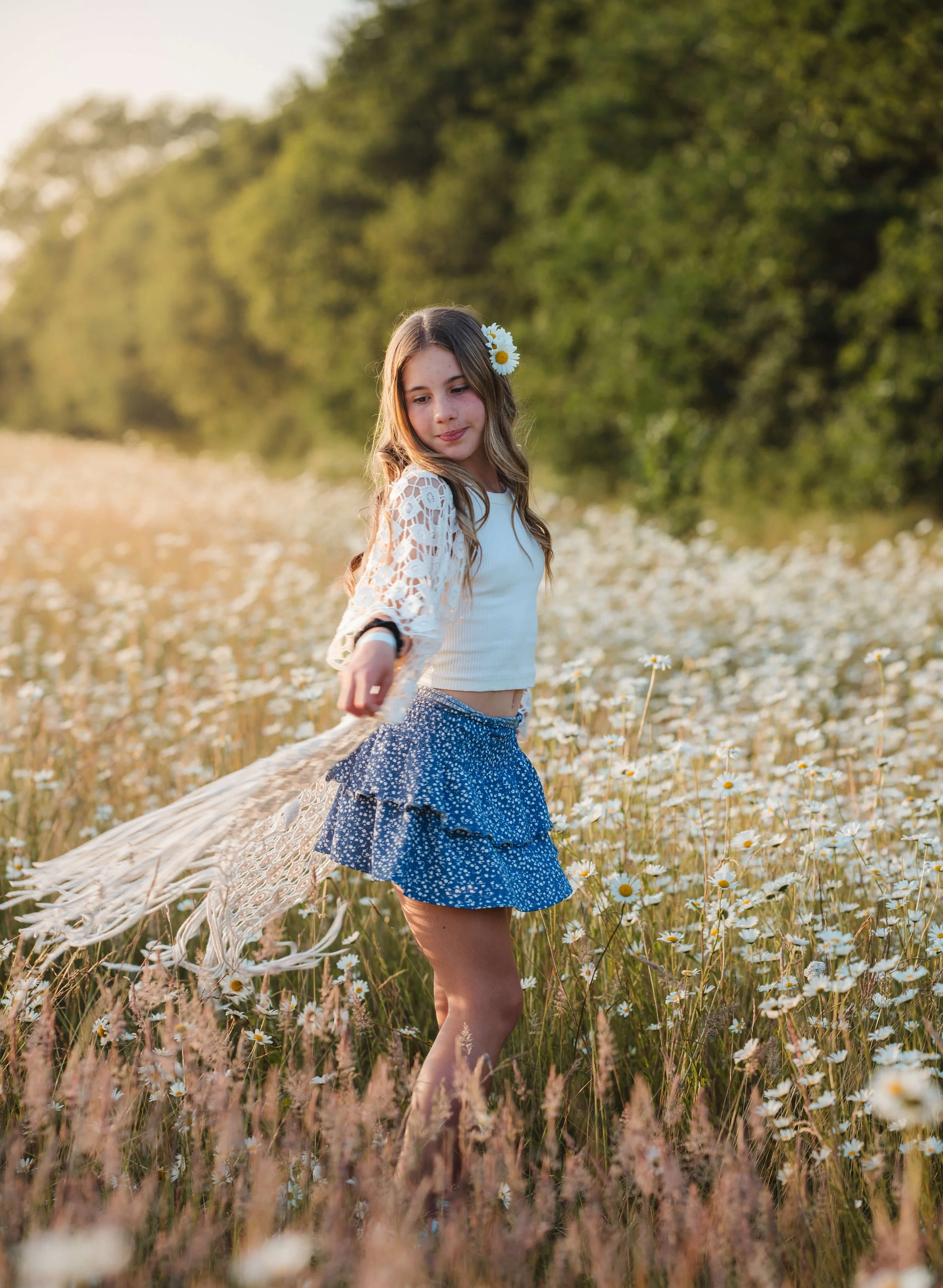 A young girl standing in a field of daisies, pointing downward, with a gentle smile and wearing a white top and blue skirt, backdropped by green trees and a sunset.
