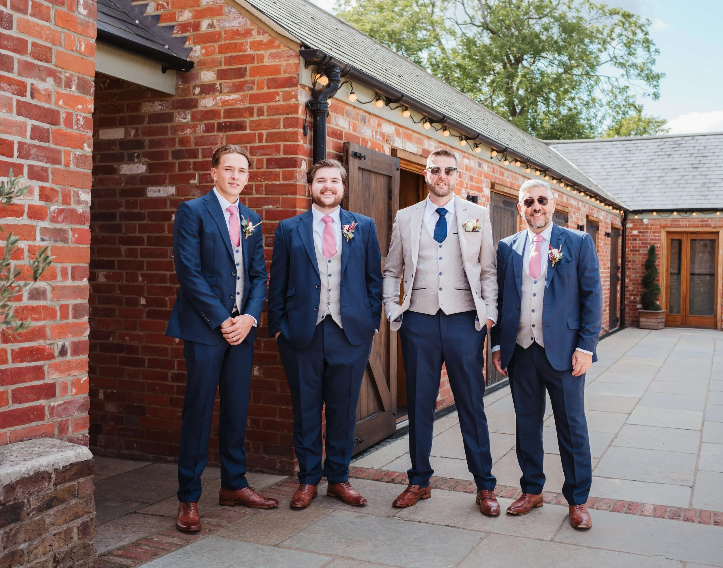 Five men dressed in suits and vests standing outside a brick building, posing for a photo. They are smiling and wearing boutonnieres.