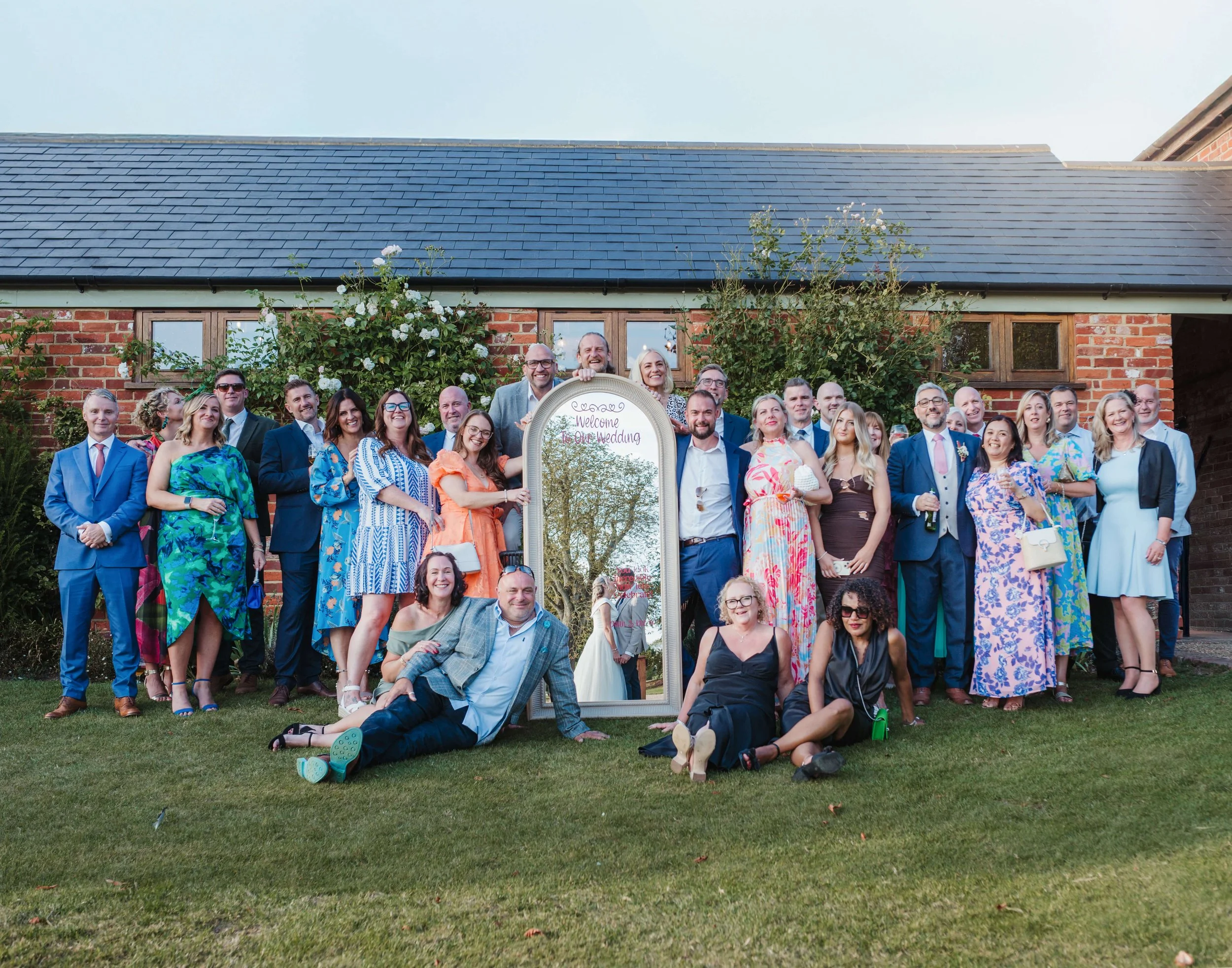 Group of wedding guests gathered outside in front of a brick building, standing around a large mirror with a handwritten sign that reads 'Welcome to Our Wedding,' reflecting trees and the wedding couple.