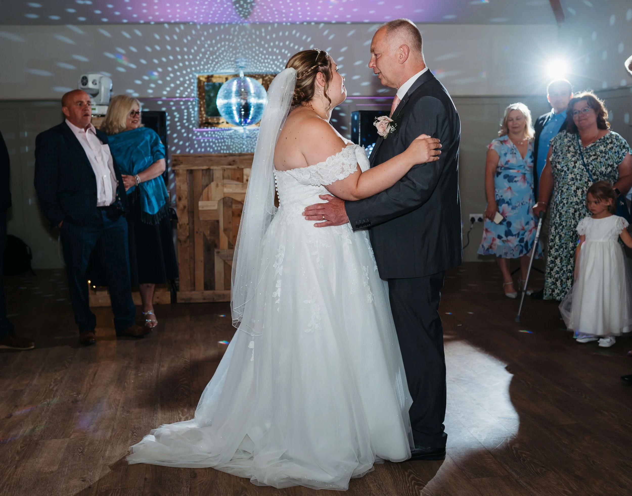 Bride and groom sharing their first dance at a wedding reception, surrounded by guests, with disco lights and a mirrored disco ball overhead.