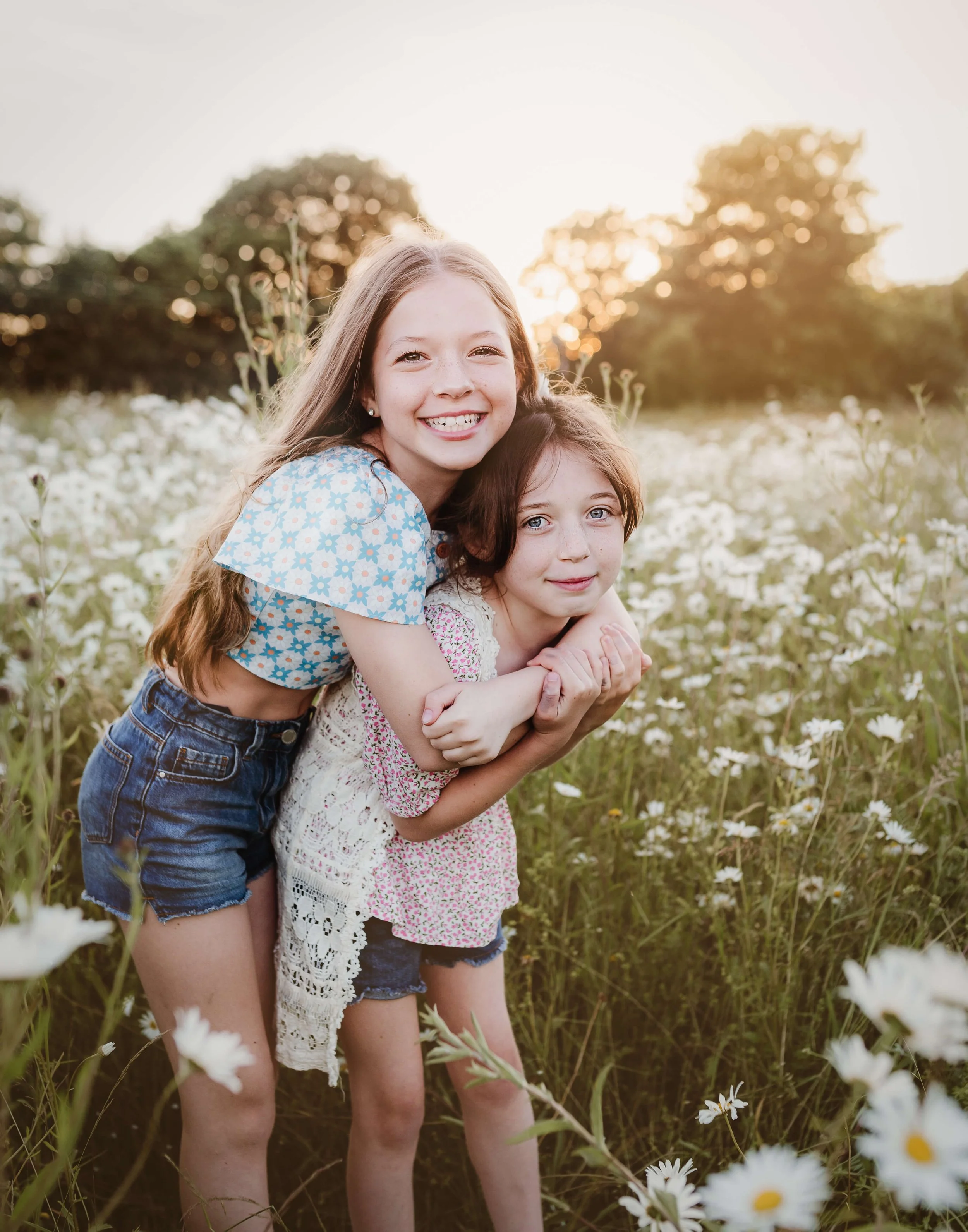 Two young girls smiling and hugging in a field of white daisies during sunset.
