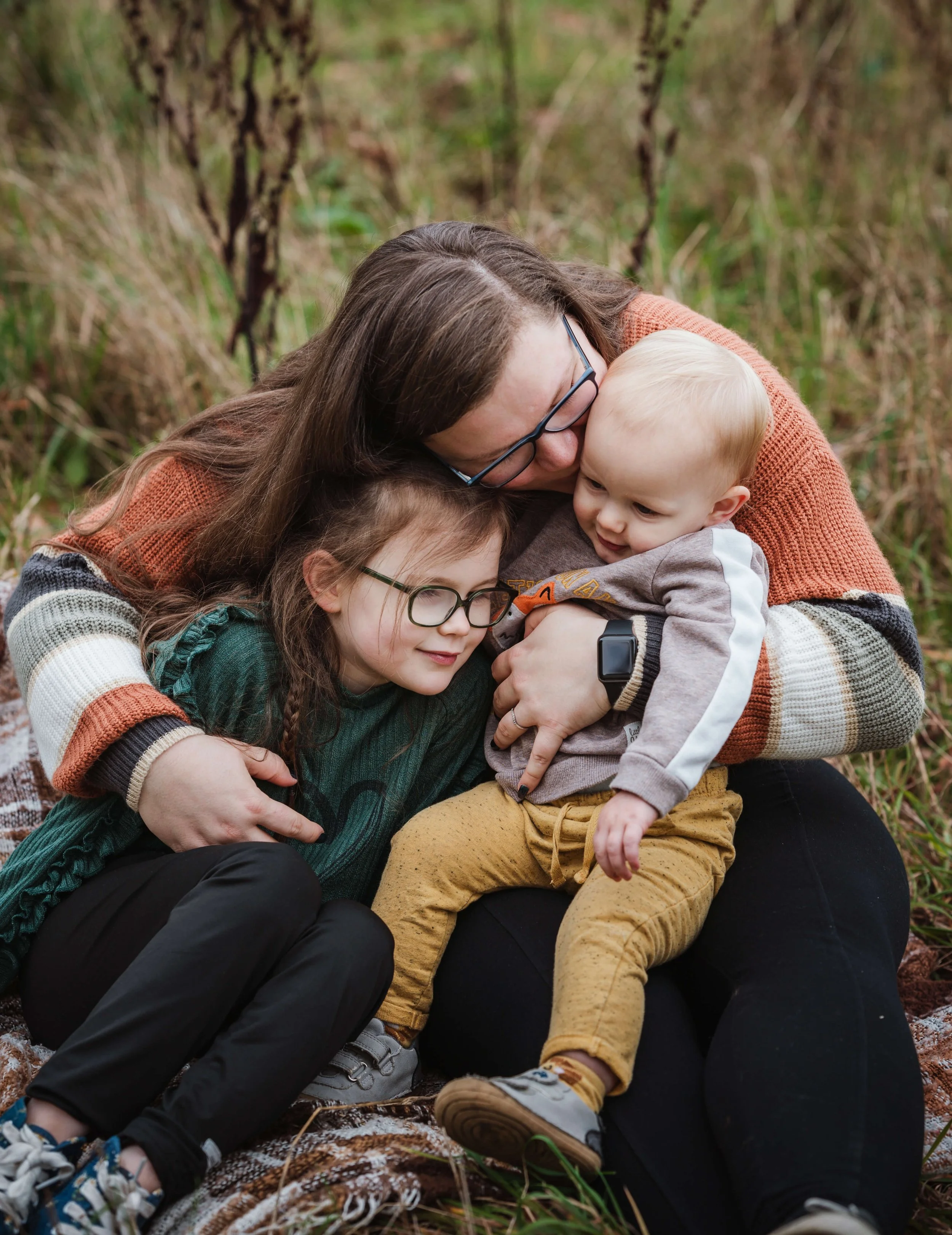 A woman with glasses hugging two children outdoors in a grassy area.