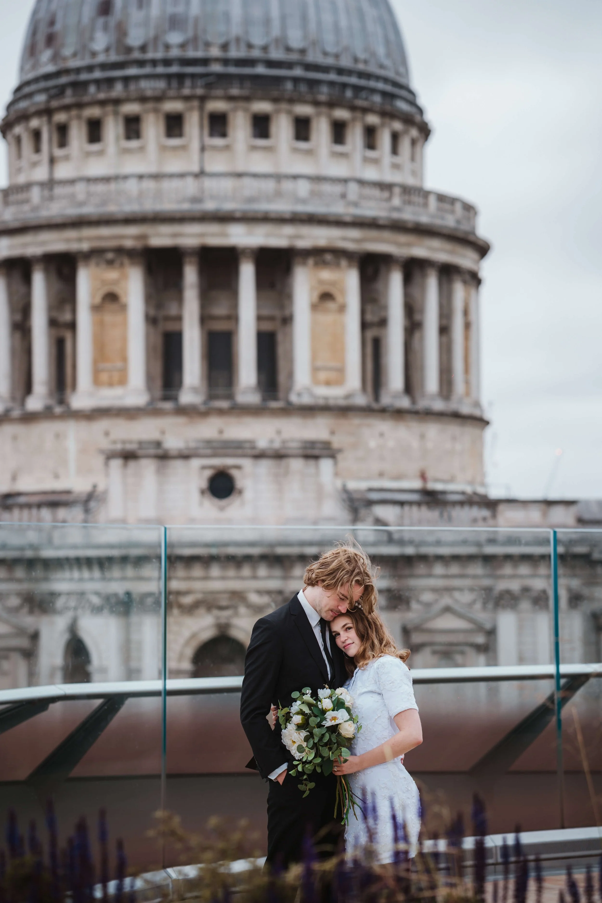A couple in wedding attire stands close together, holding a bouquet of white flowers, with the historic St. Paul's Cathedral in London in the background.