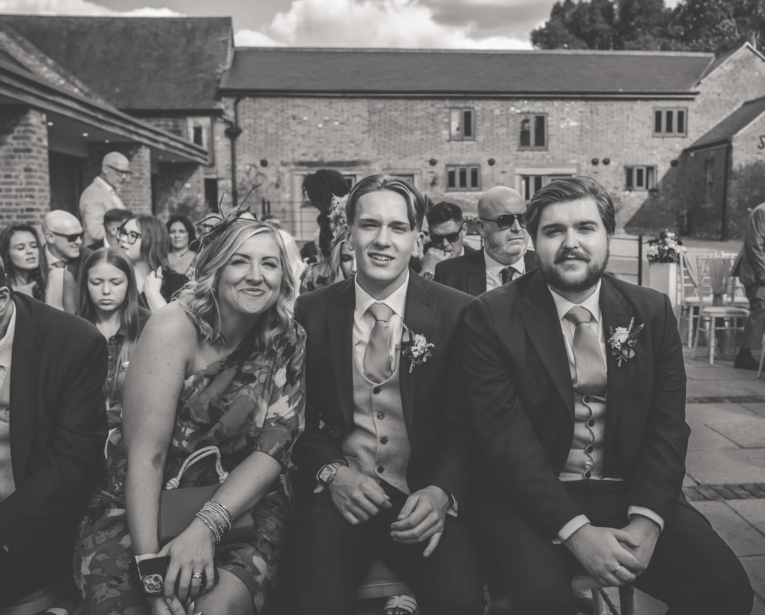 Black and white photo of a group of people at an outdoor wedding ceremony, including two men in suits and a woman in a floral dress sitting in the front row.
