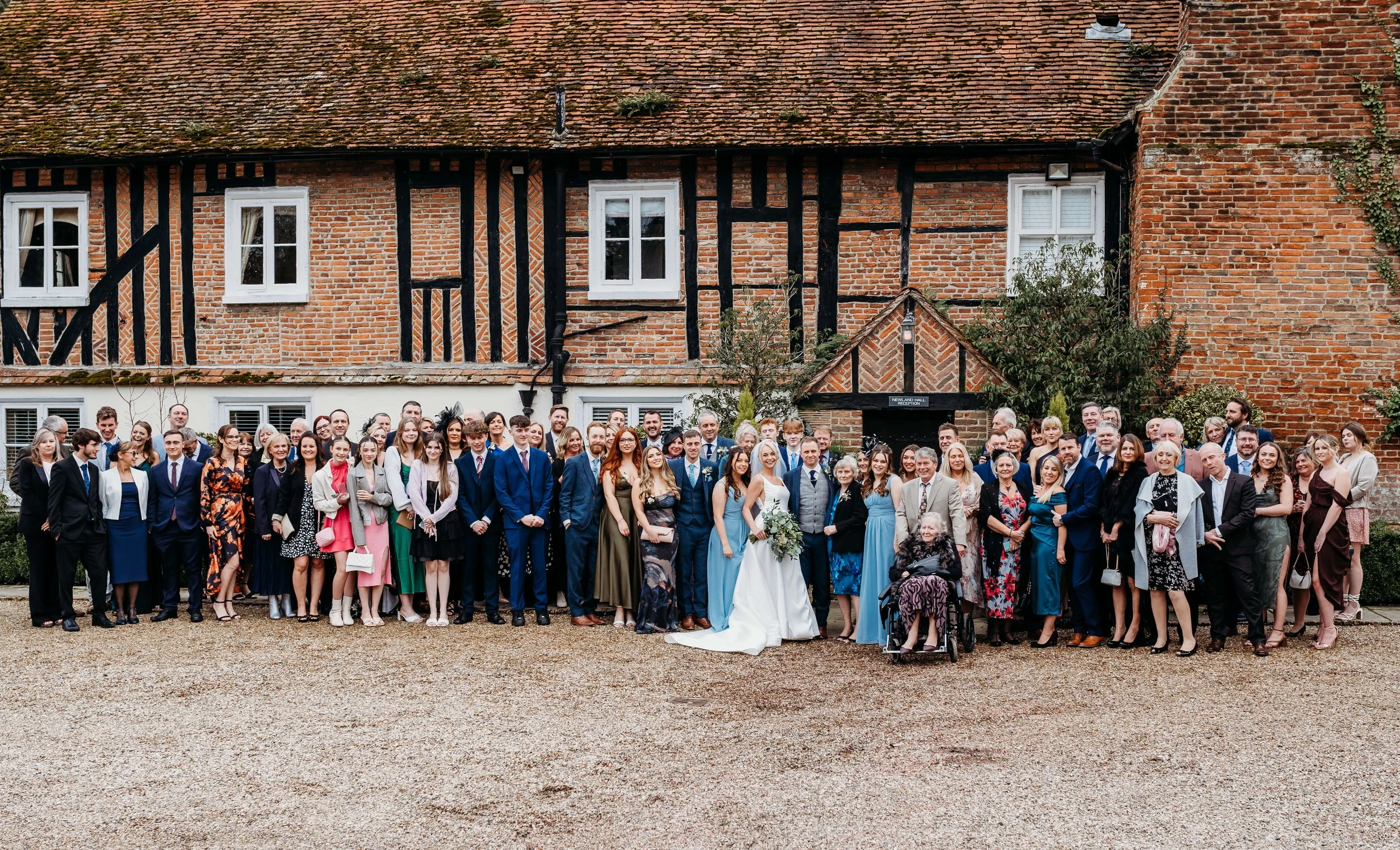 A large group of people, dressed in formal attire, gathered outside in front of a brick and timber-framed building for a celebration, likely a wedding.