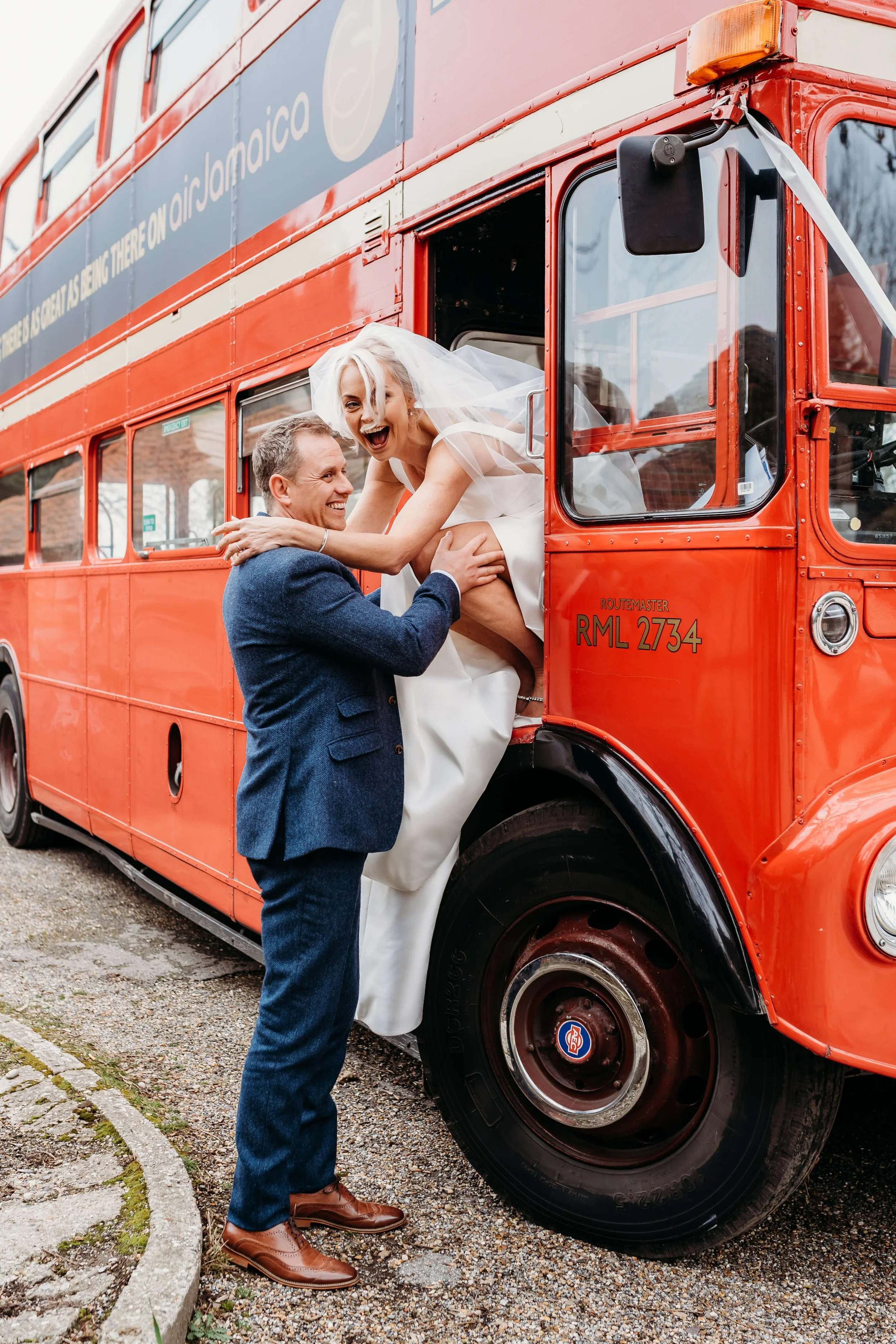 A man dressed in a navy blazer and brown shoes is lifting a woman in a white wedding dress and veil out of the doorway of a red double-decker bus. The woman is smiling and appears excited.