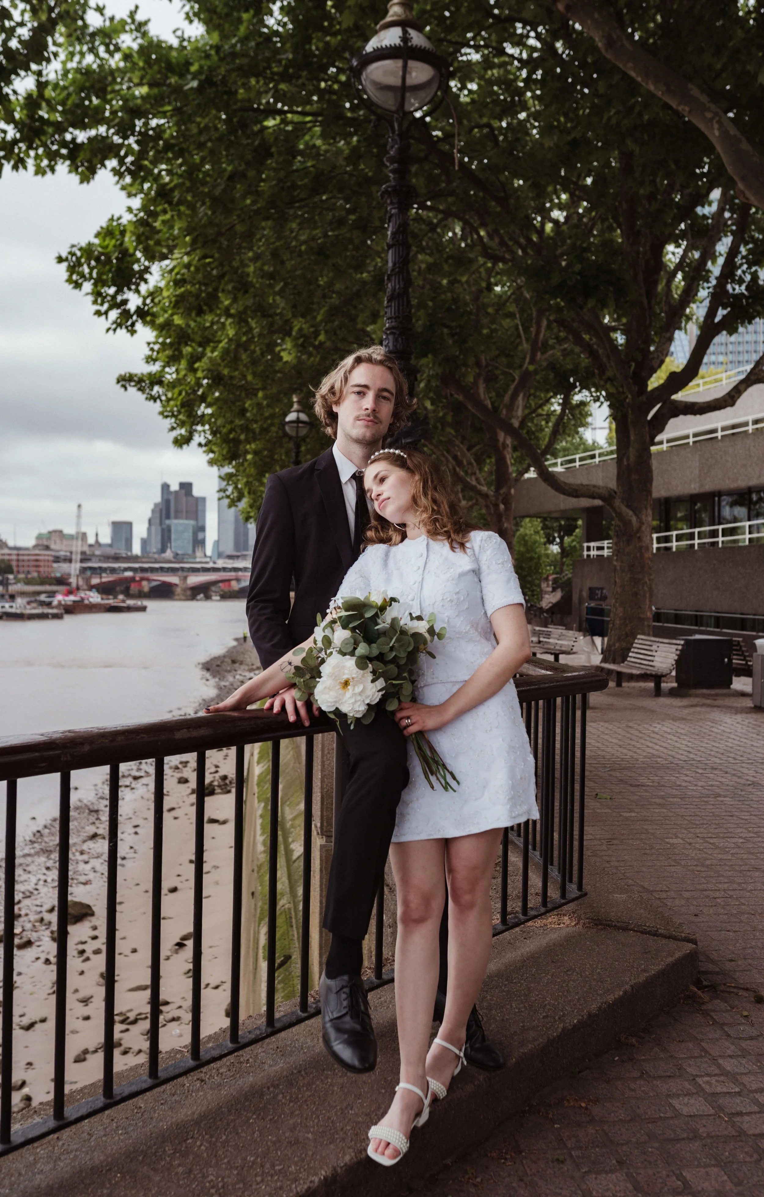 A couple dressed in wedding attire standing on a riverside promenade, with buildings and a bridge in the background, under leafy trees.
