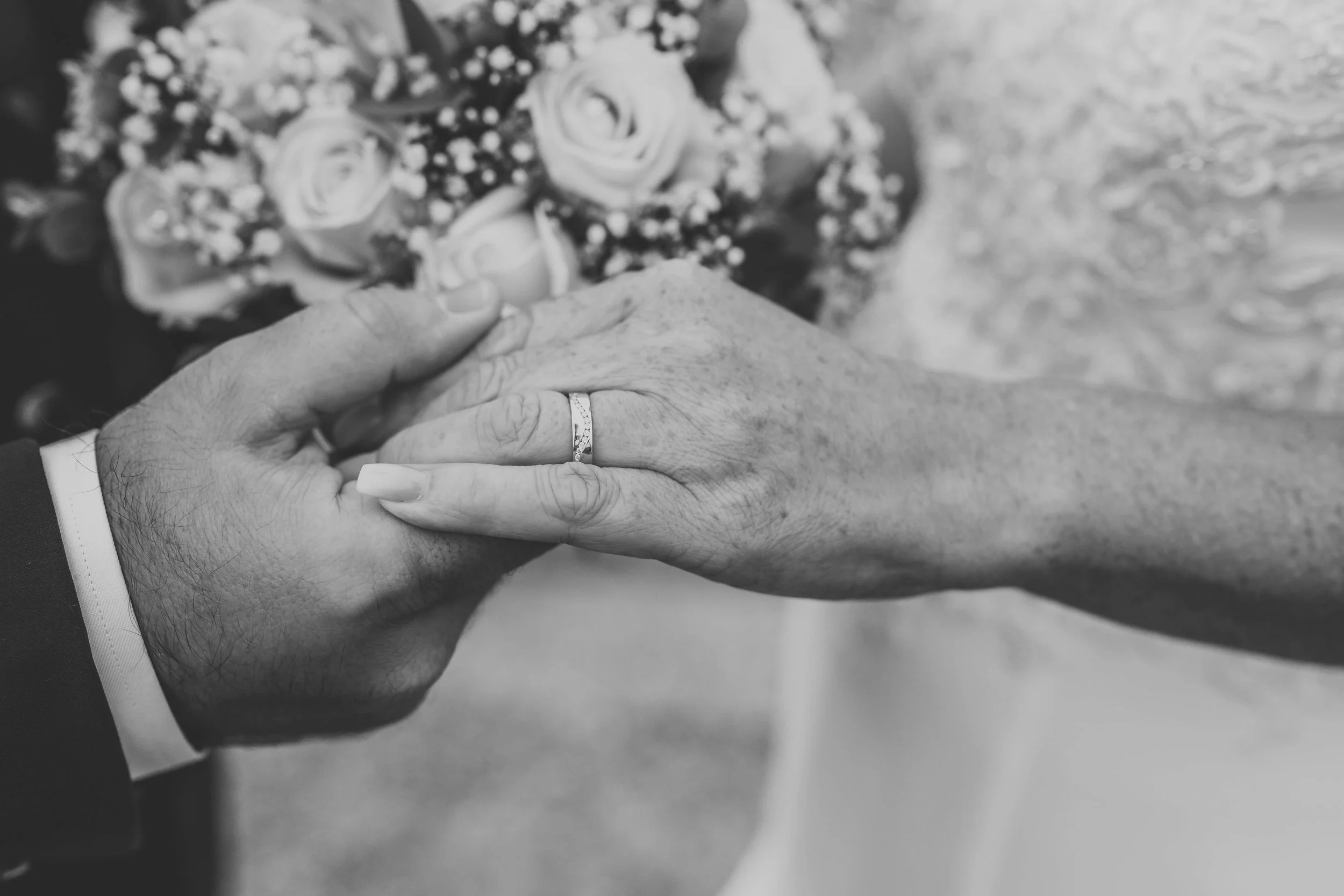 Close-up of a groom's hand holding a bride's hand, showing wedding rings, with a bridal bouquet in the background.