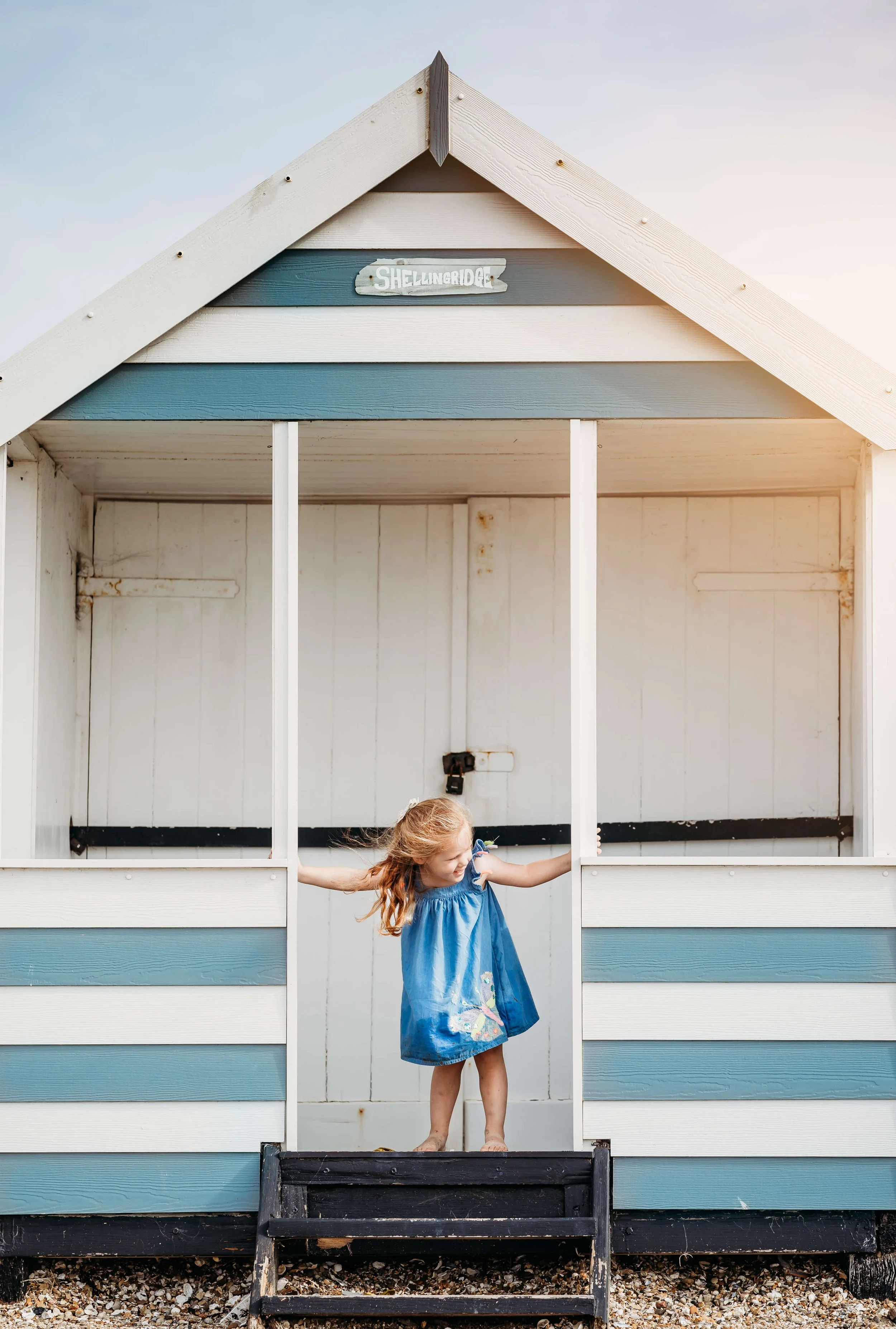 A young girl in a blue dress standing on the steps of a small, empty beach house with a sign labeled 'Shellingridge'.