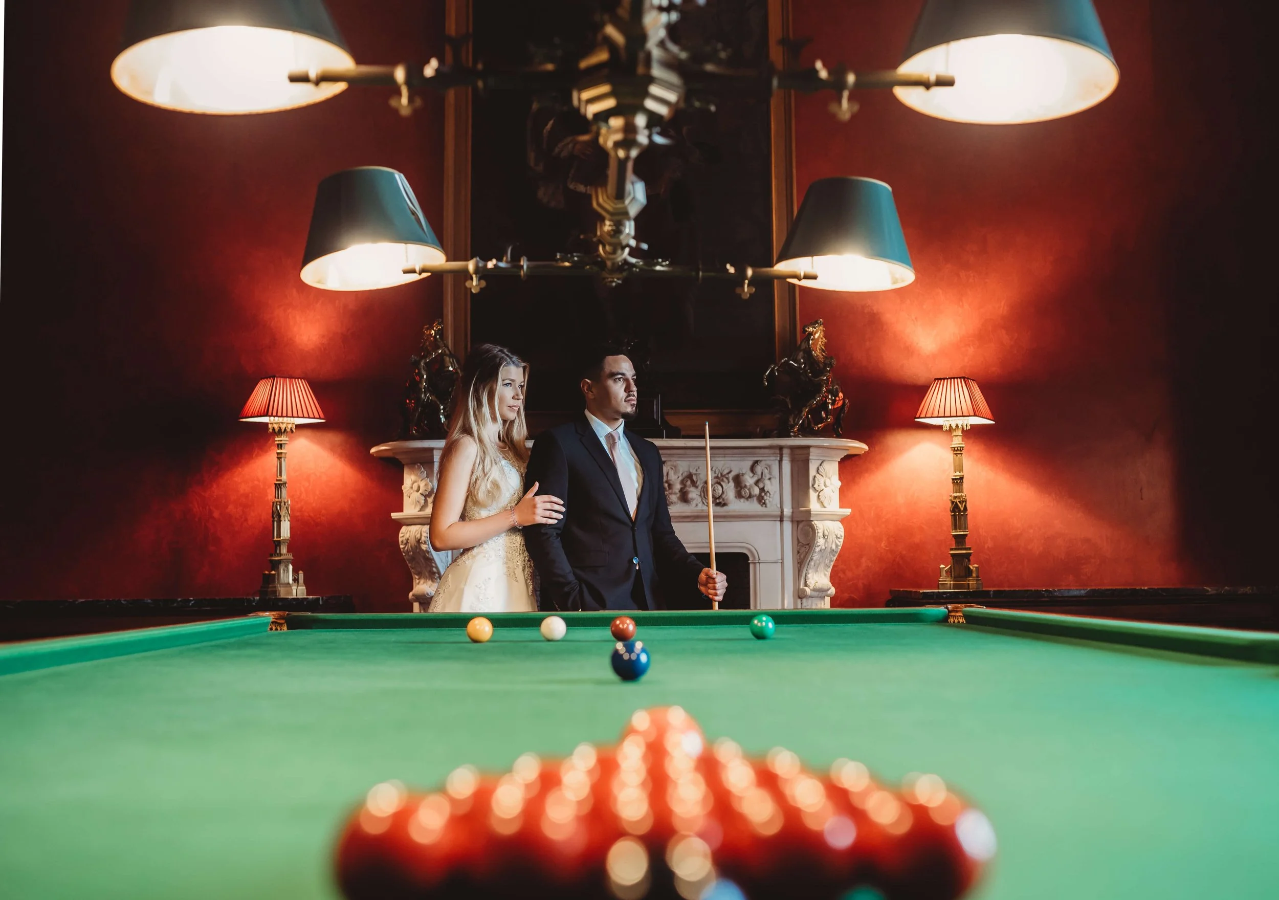 A man and woman in formal attire standing by a pool table in a luxurious room, with a fireplace and ornate decorations in the background.