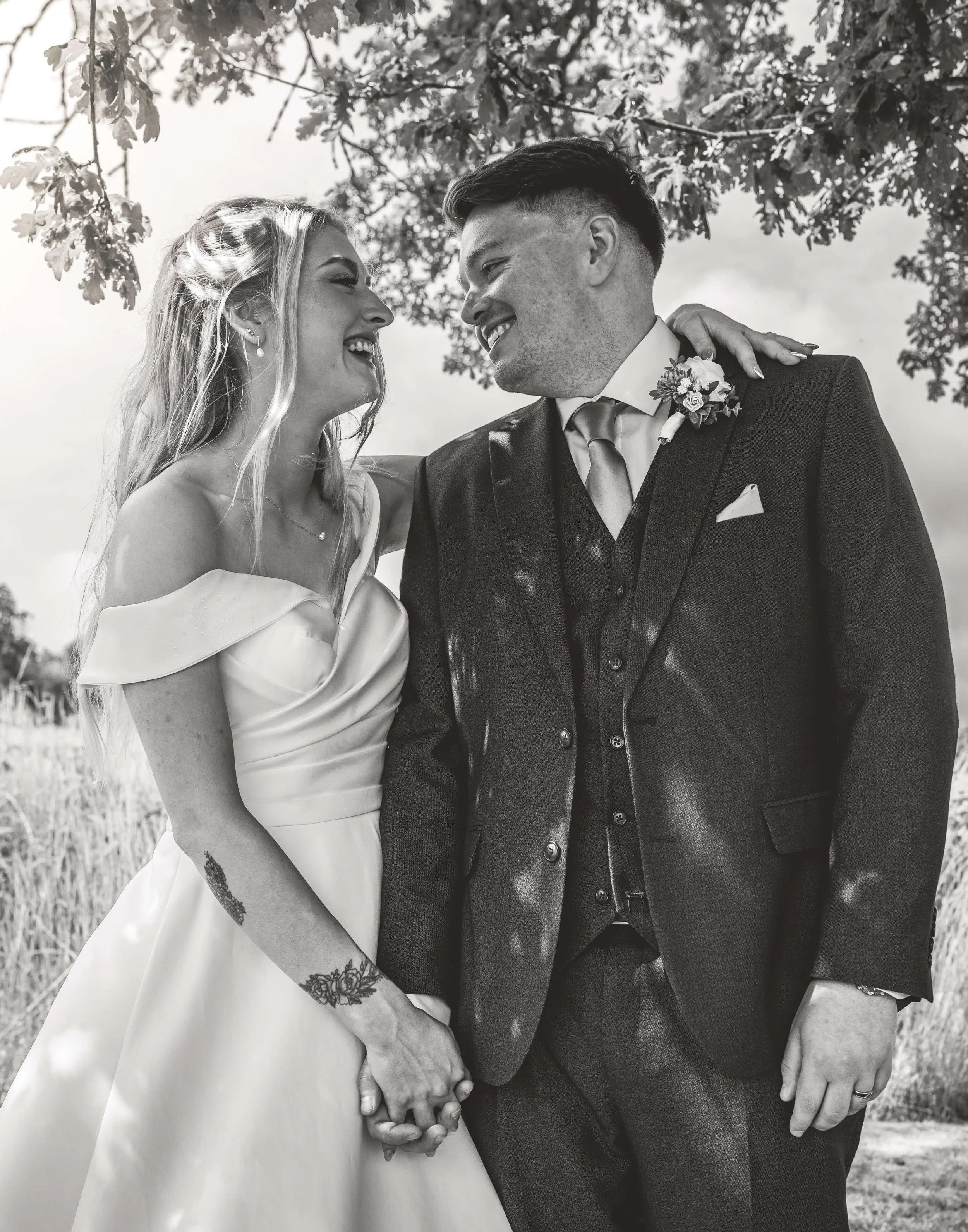Black-and-white photo of a wedding couple smiling at each other, holding hands outdoors under a tree.
