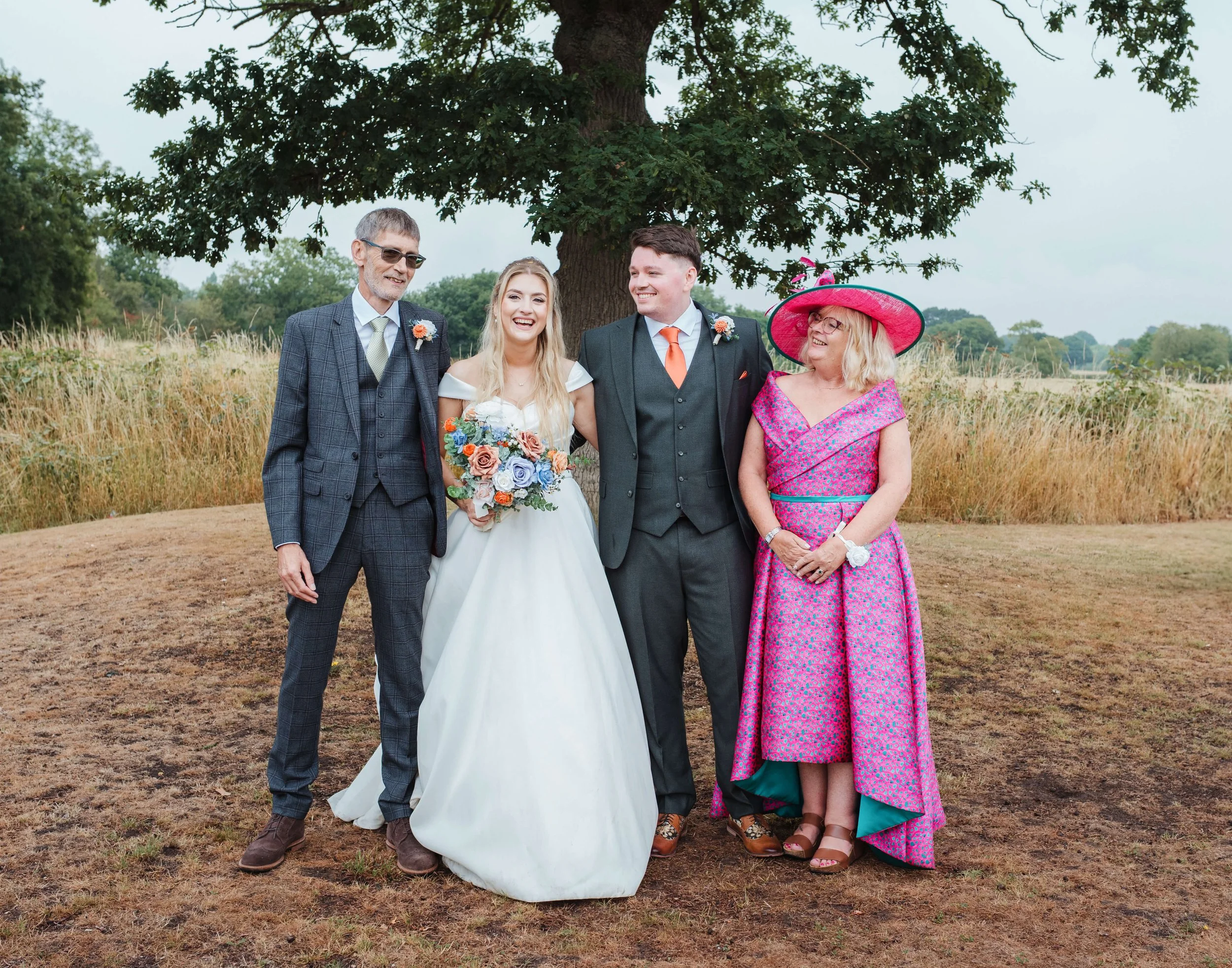 A group of five people at a wedding outdoors, including a bride in a white gown holding a colorful bouquet, a groom in a dark suit with an orange tie, an older man in a checkered suit, and an older woman in a bright pink dress with a large pink hat, 